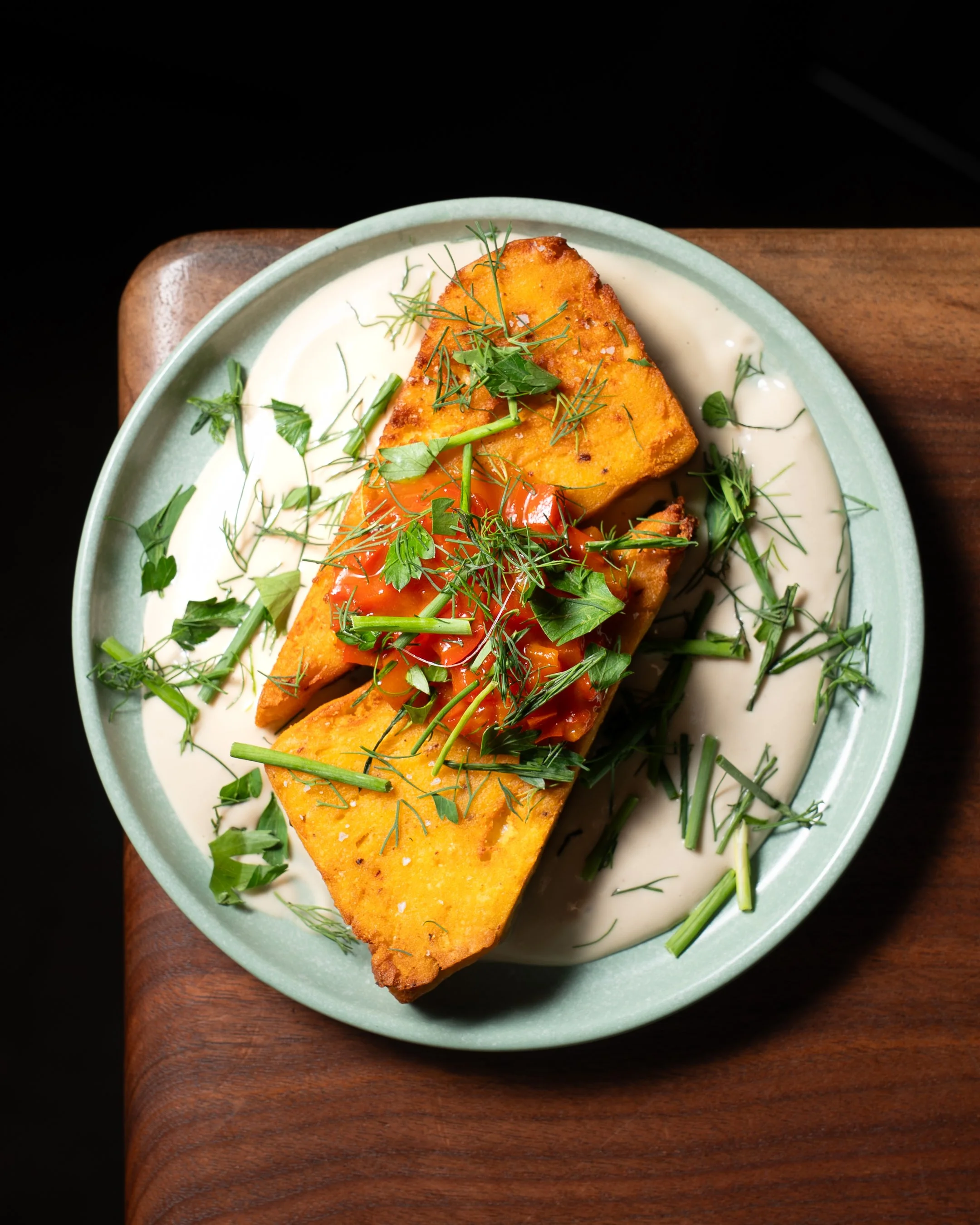 A plate with sections of fried tofu topped with fresh green herbs, drizzled with red sauce, and served on a bed of white sauce or dressing, all on a green ceramic dish resting on a wooden surface.