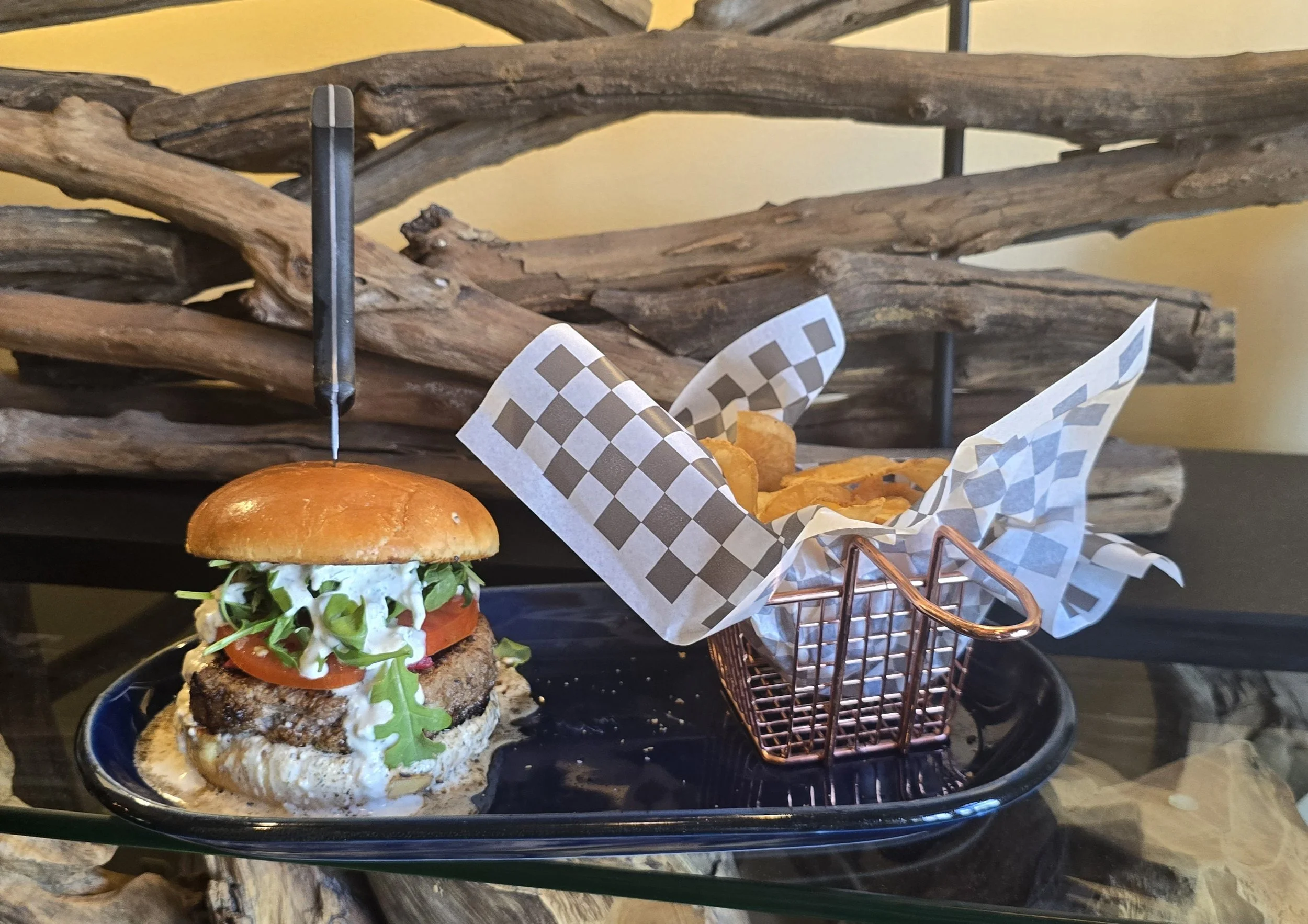 A gourmet burger with lettuce, tomato, onion, and creamy sauce on a toasted bun, served on a black tray with a small copper basket of French fries, placed on a dark glass table against a rustic wooden backdrop.