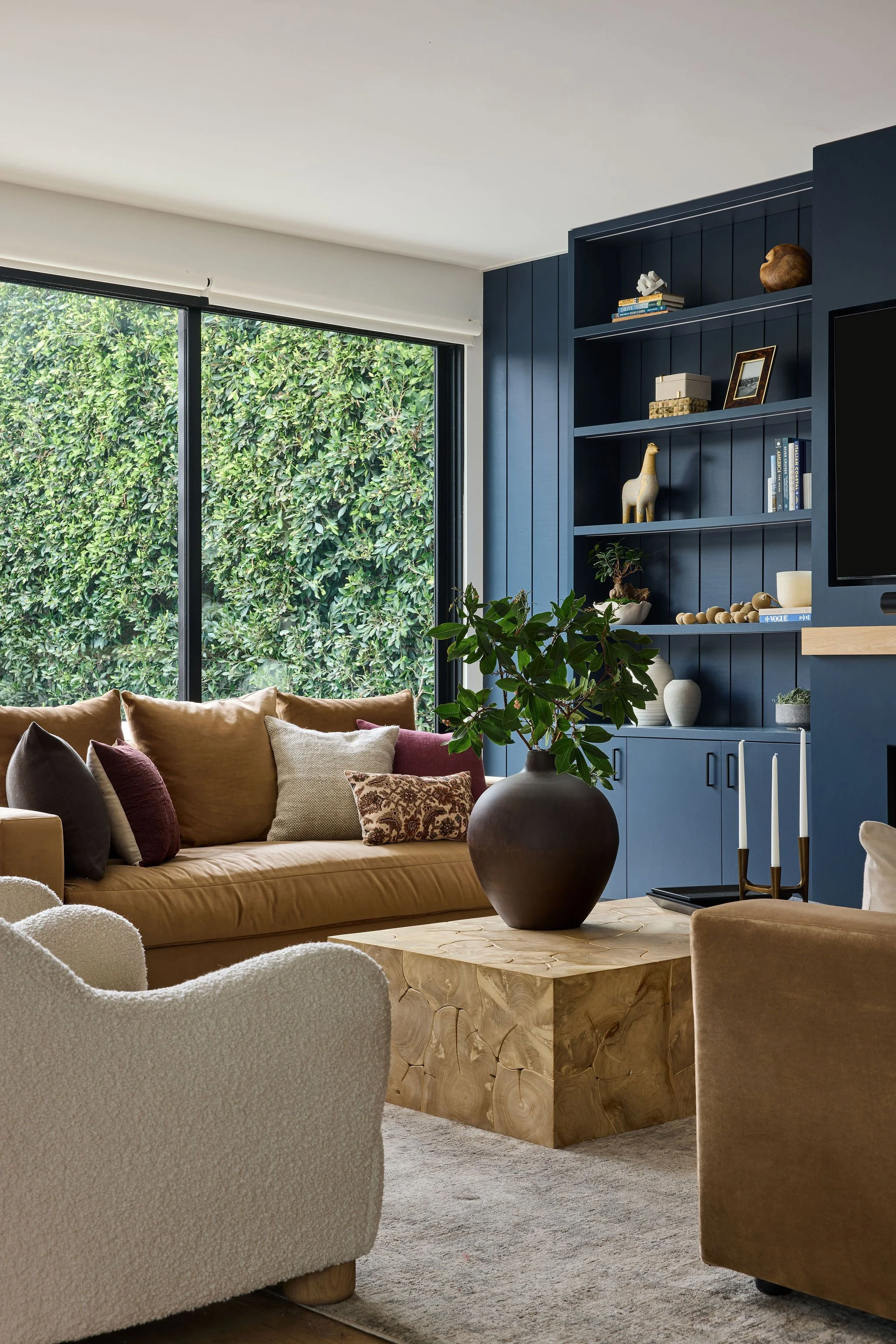 Living room with beige, white, and dark blue couches, a wooden coffee table, a large vase with green plants, a wall with shelves and decor, and a large window with green foliage outside.
