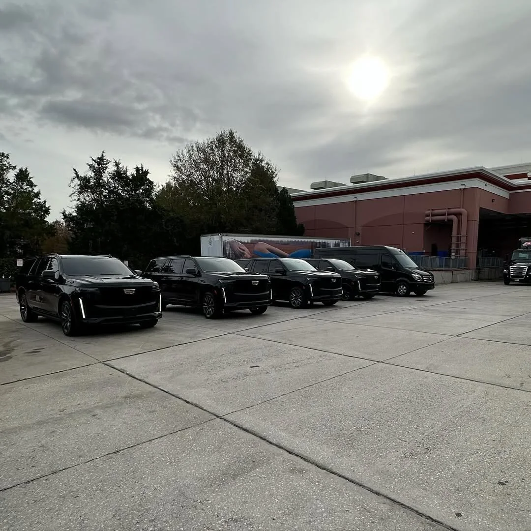 A parking lot with five black SUVs parked in a row, a black van, and a semi-truck at a warehouse or distribution center on a cloudy day with the sun partially visible through the clouds.
