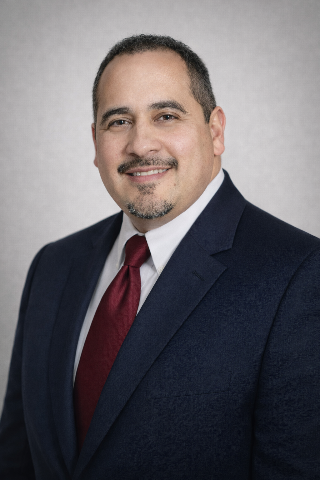 A professional headshot of a man in a navy blue suit, white shirt, and red tie, smiling against a grey background.