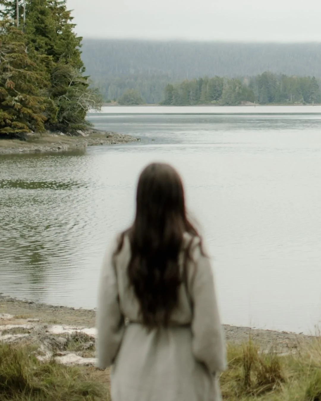 A woman with long dark hair stands facing a lake, with trees on the hornby island shoreline relaxing after her massage treatment.
