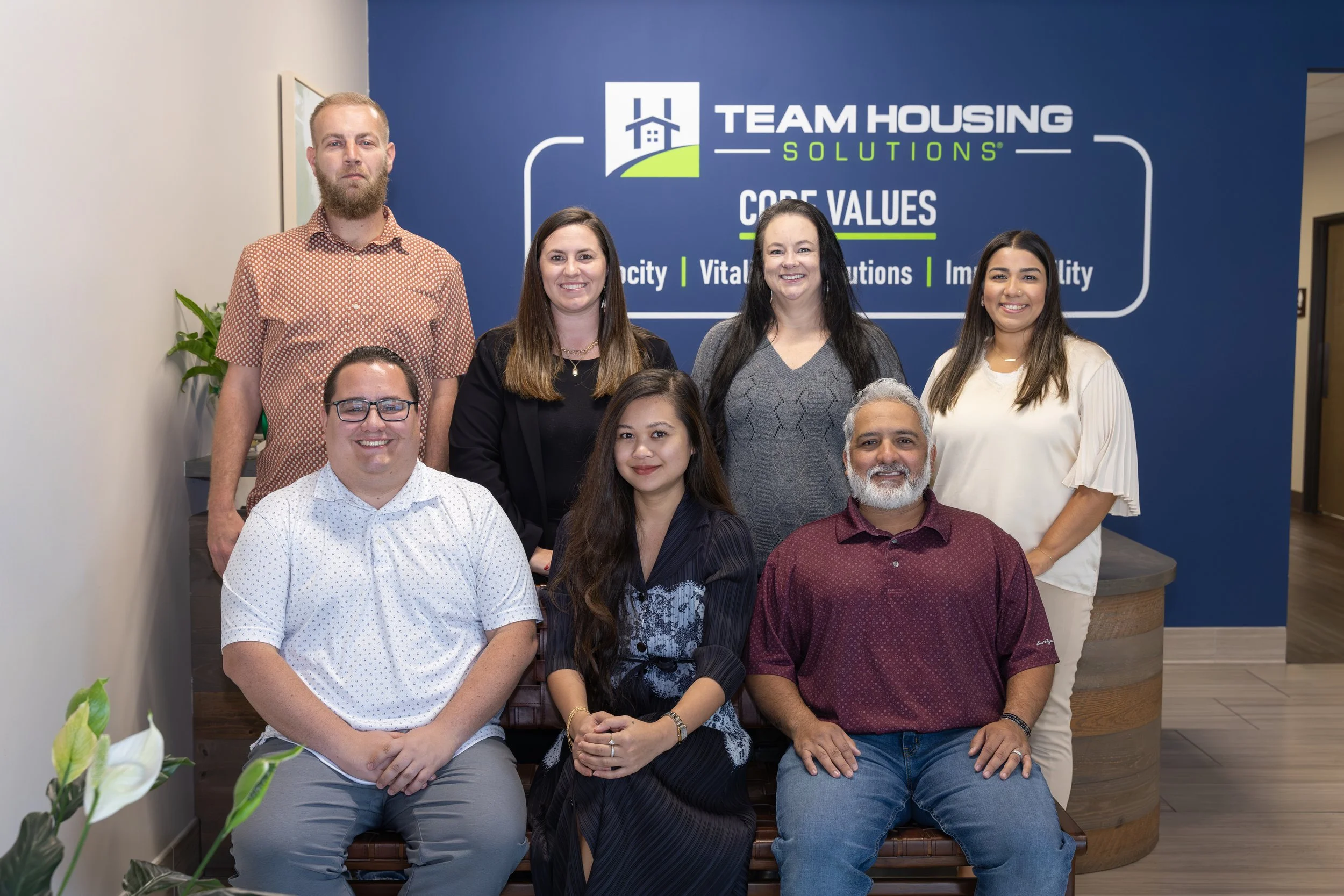 A group of nine diverse team members posing in front of a blue wall with the team logo and core values of Team Housing Solutions, smiling for the camera.