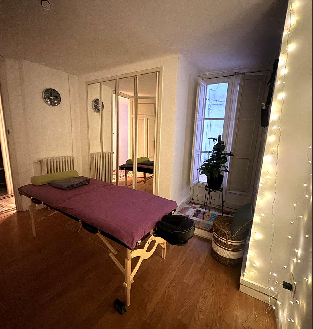 A room with a massage table covered with a pink sheet and a gray towel, a mirror closet, a window with open shutters letting in natural light, a potted plant on a stand, a woven basket, and string lights on the wall.