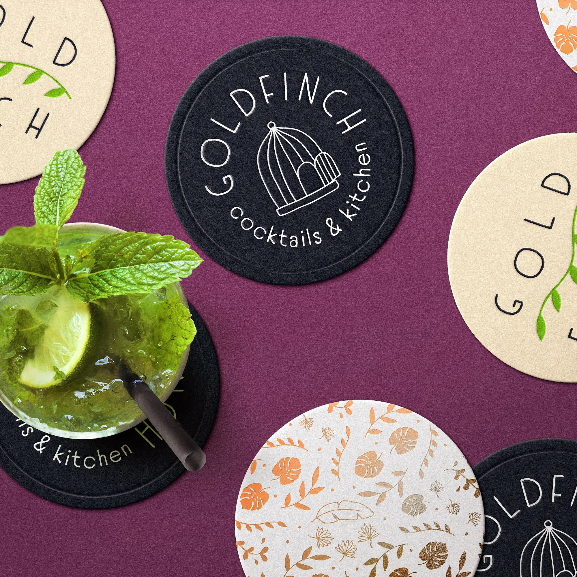 Black coasters on a purple table top with the hand-drawn bird cage logo for Goldfinch Cocktails and Kitchen on them.