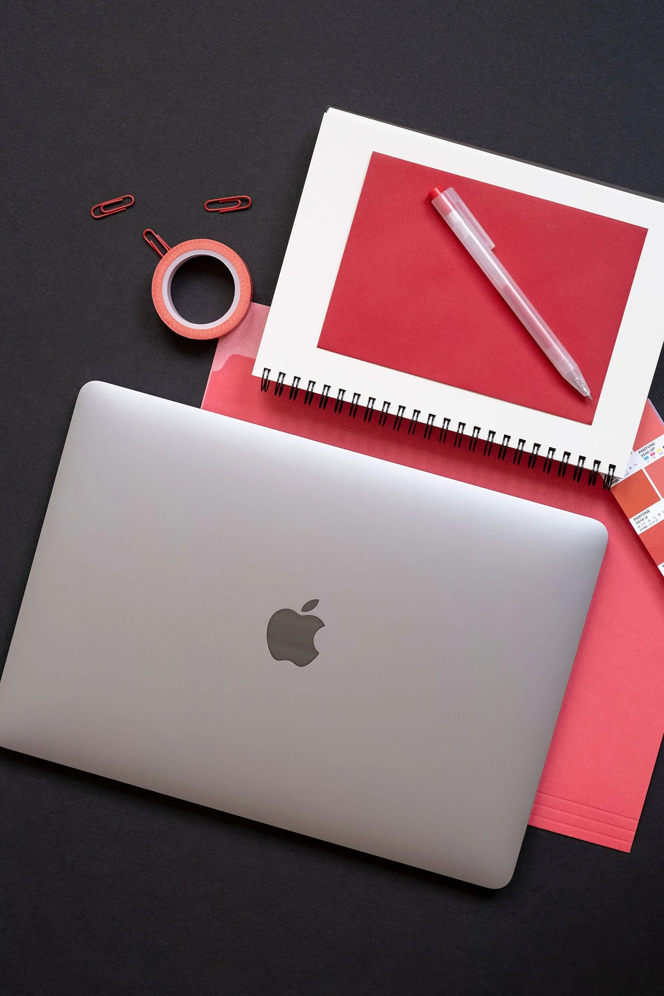 Top-down view of an Apple laptop on top of a black table top with a red file folder, spiral notebook, red pen, red envelope, red tape, and Pantone swatch book open to red swatches.