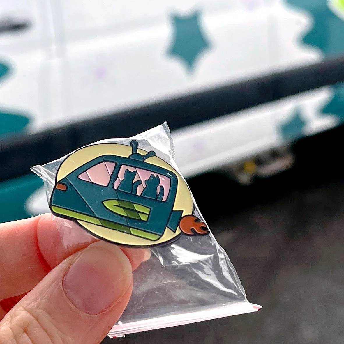 Fingers holding an enamel pin of a mobile veterinary van that looks like a spaceship with a wing on the side and a flame jetting off the back, and a dog and cat in the window