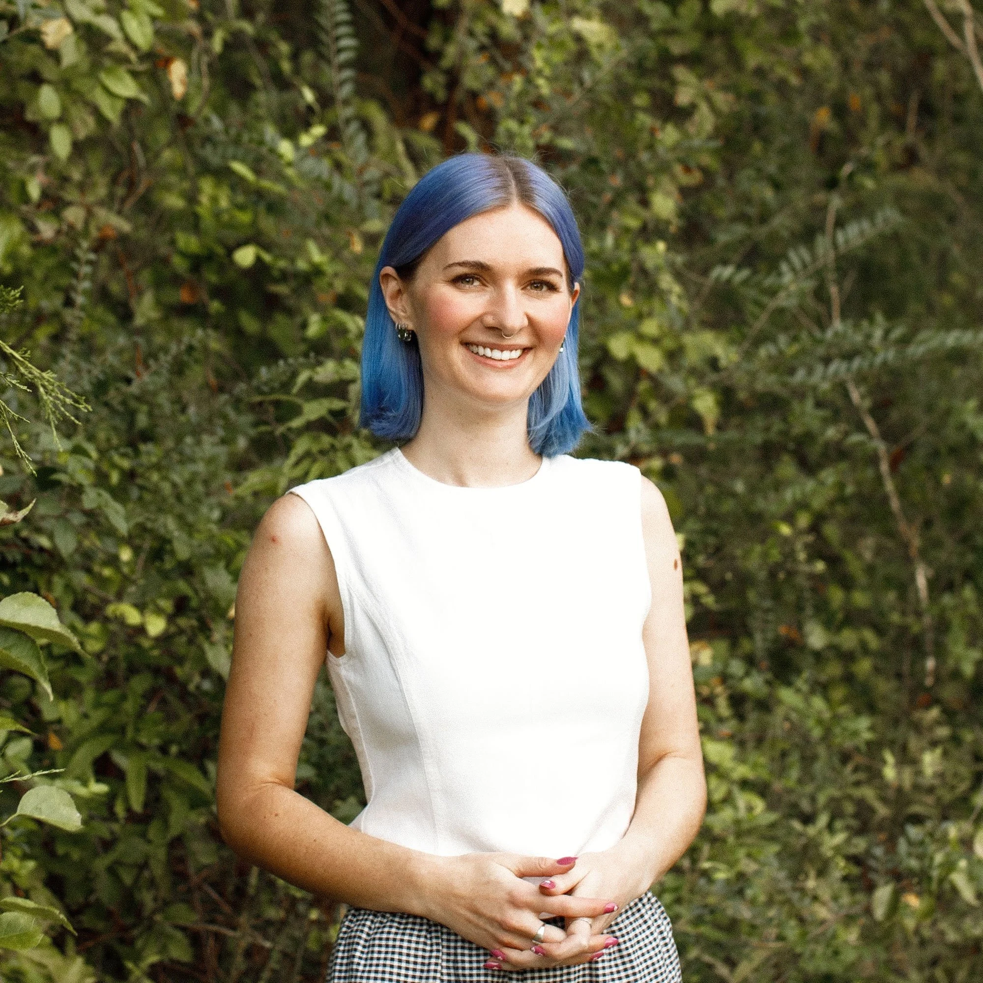 Cass, Shopify developer, standing and smiling with her hands folded in front of her and green foliage behind her.