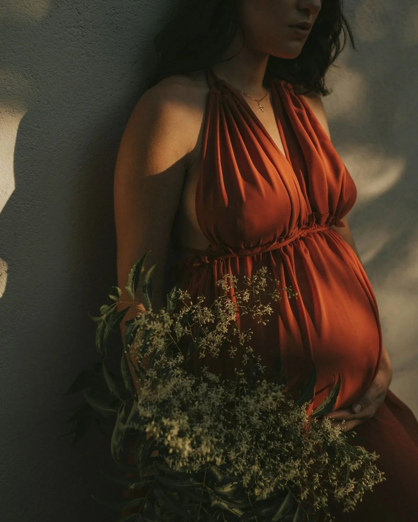 An expecting mother wearing a rust-colored dress with thin straps standing against a textured wall, holding some green foliage and small white flowers.