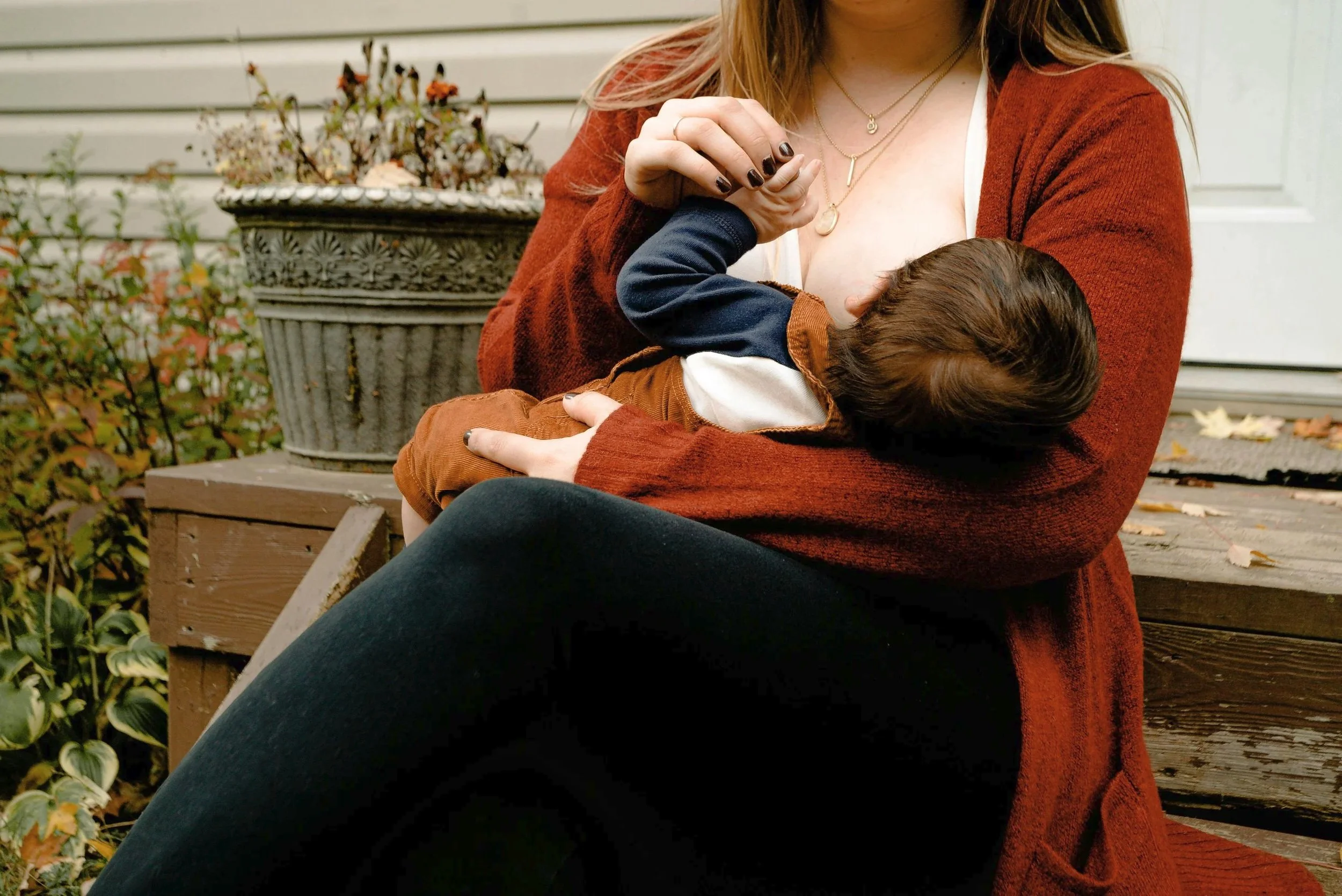 A woman sitting on a wooden bench outdoors, holding and nursing her baby in her arms, with autumn leaves on the ground and potted plants nearby.
