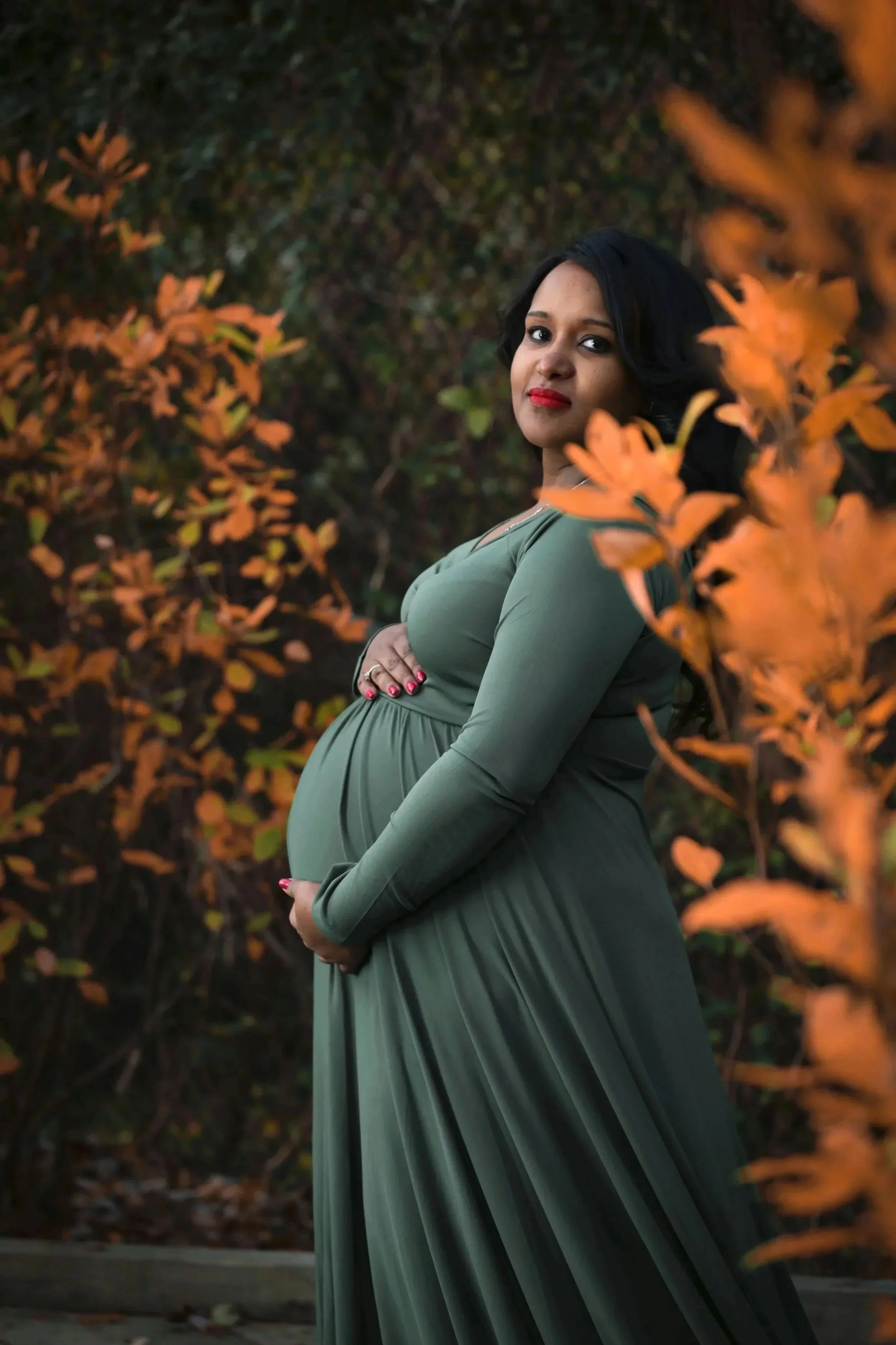 A pregnant woman in a long, green dress standing amidst orange autumn leaves, with her arms crossed over her belly, smiling softly.