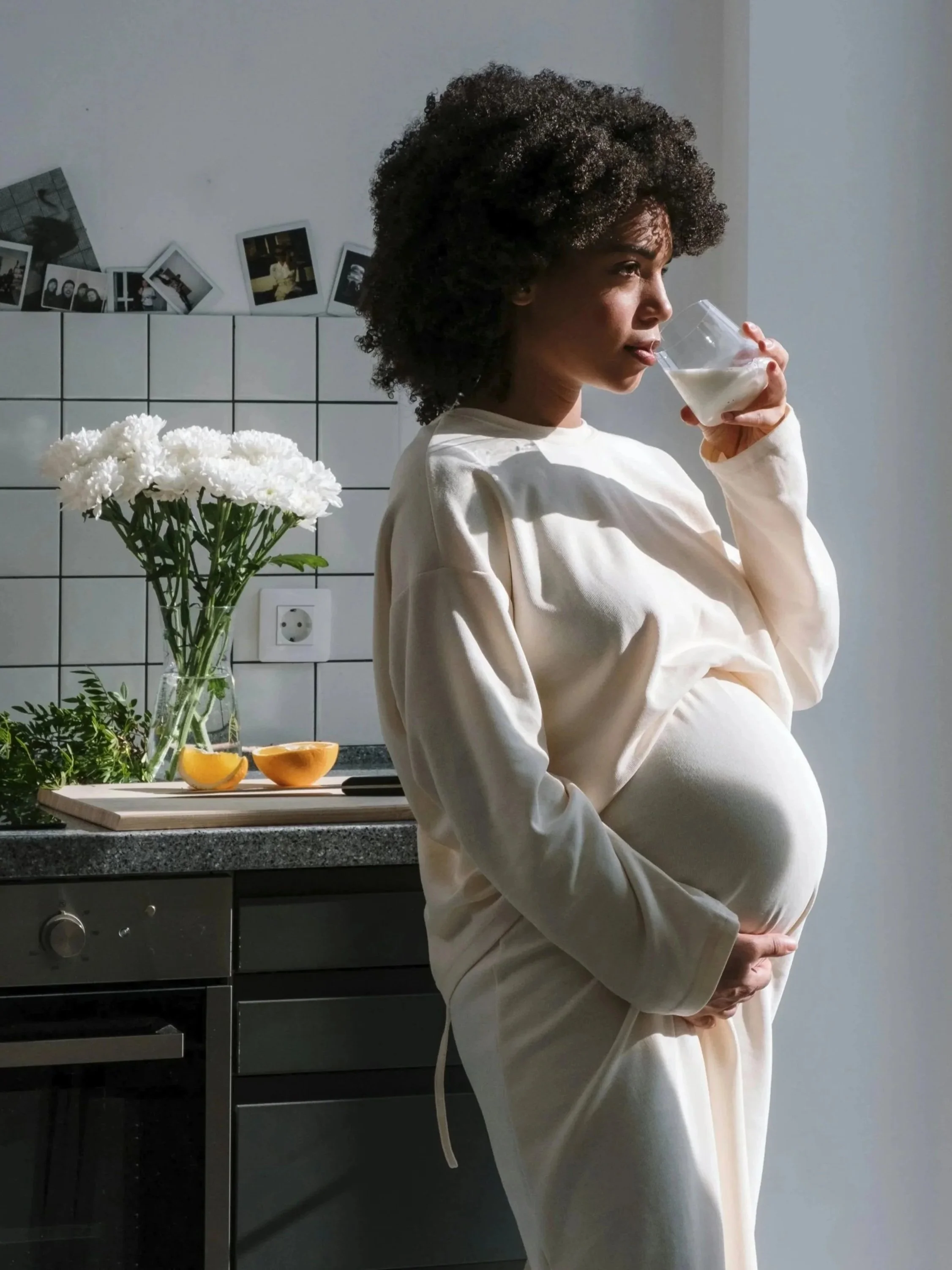 A pregnant woman with curly hair in a cream-colored dress is standing in a kitchen, drinking from a clear glass of milk. There are white flowers in a vase, half-cut oranges, and herbs on the countertop. The wall behind has a collection of photographs, and an electrical outlet is visible.
