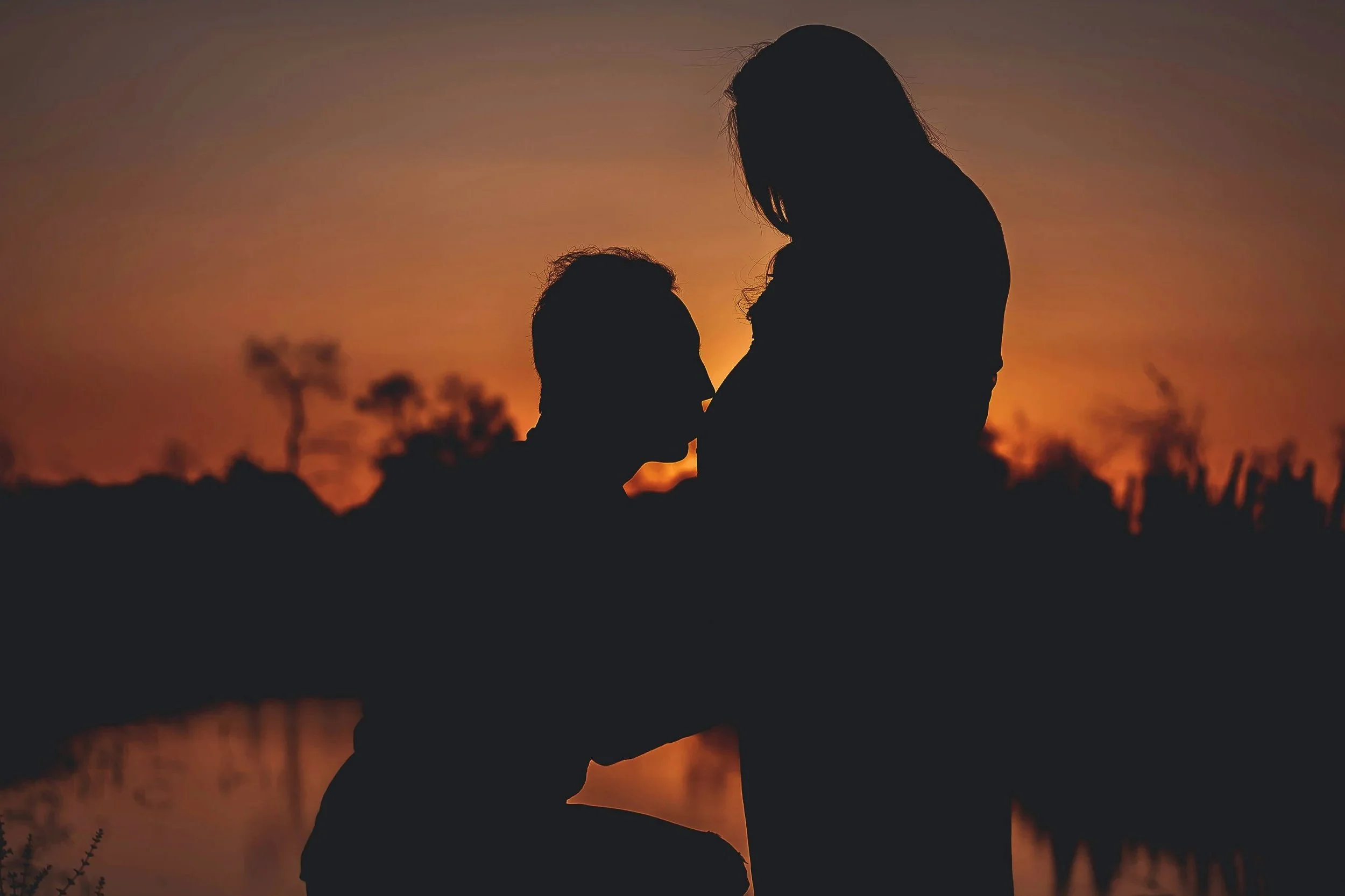 Silhouettes of a pregnant woman and her partner kissing her belly during sunset.