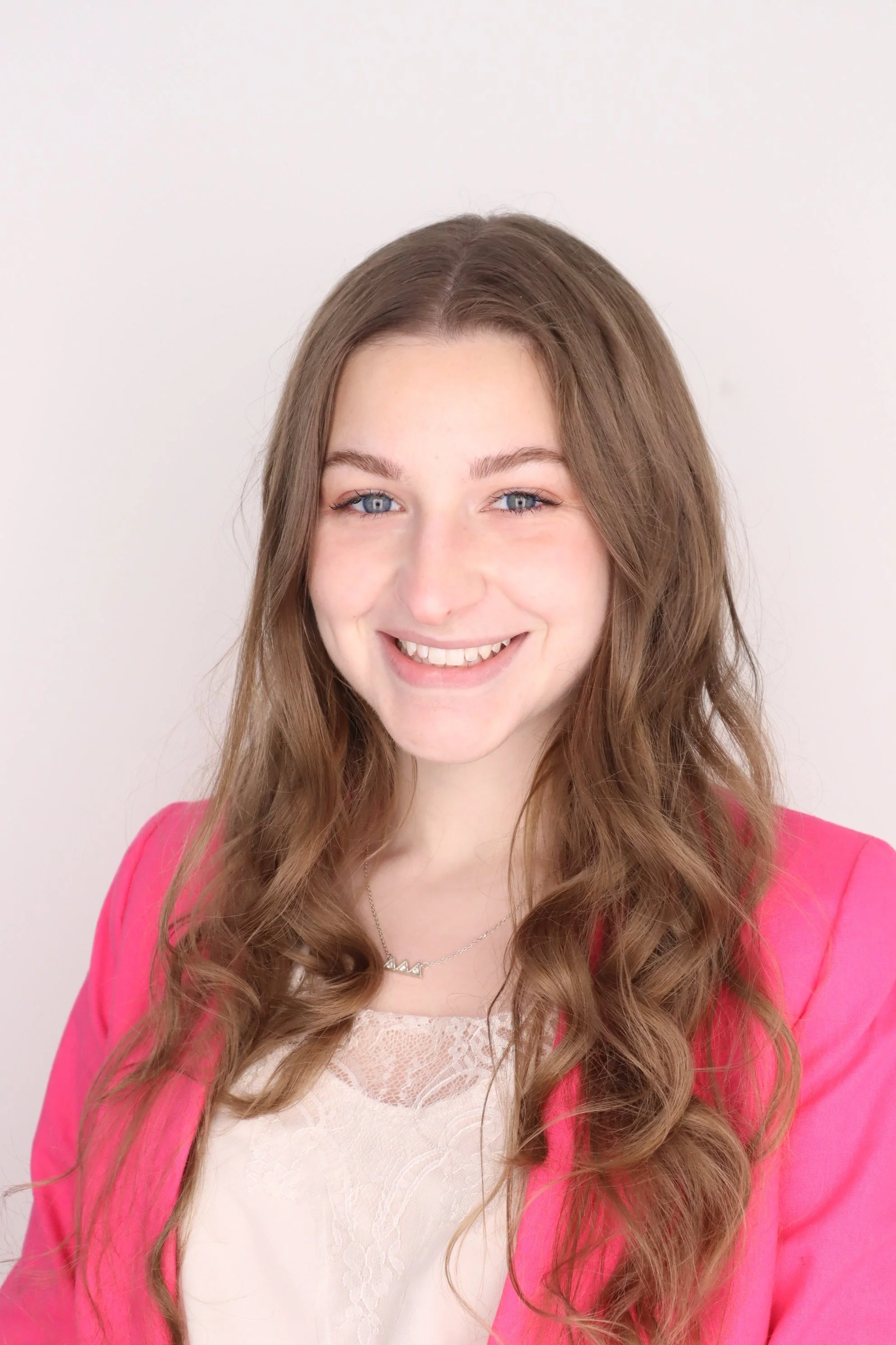 A young woman with glasses and braids smiling outdoors in front of a leafy tree and a brick wall, wearing a white shirt and a light-colored cardigan.