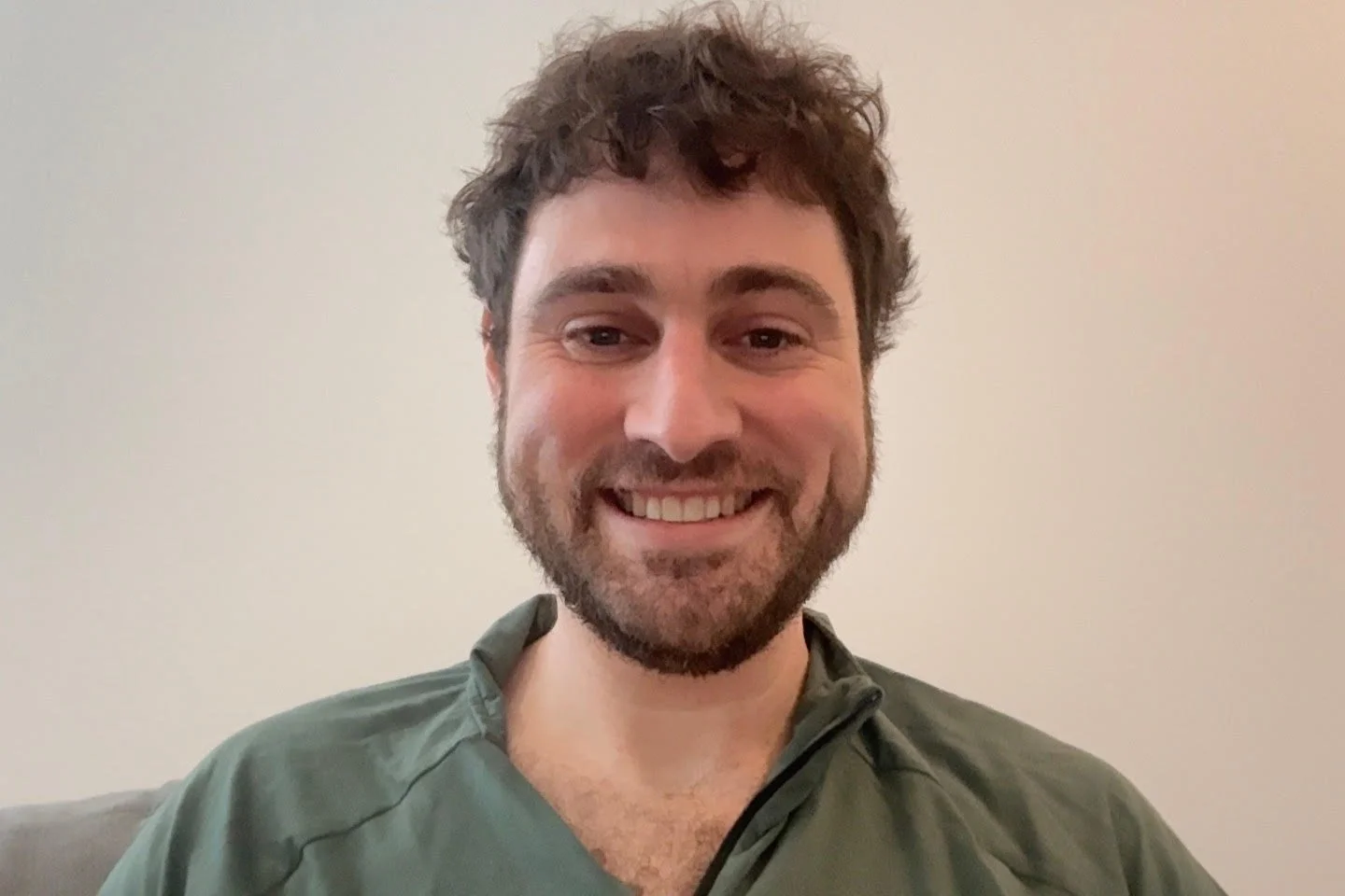 A smiling man with dark, curly hair and a beard, wearing a green zip-up shirt, sitting against a plain, light-colored wall.