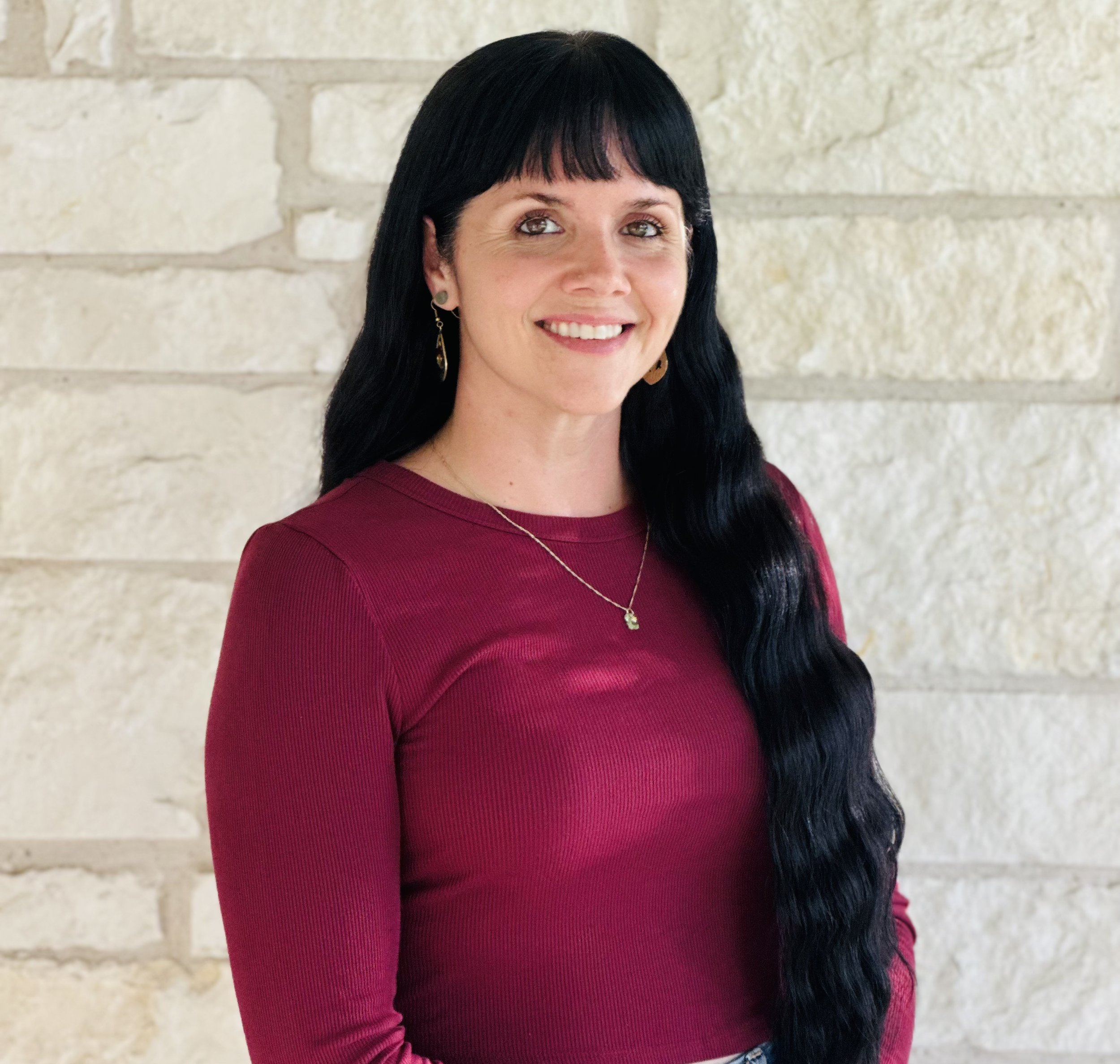 Woman with long black hair, wearing a maroon long-sleeve top, jewelry, and smiling, standing in front of a beige brick wall.