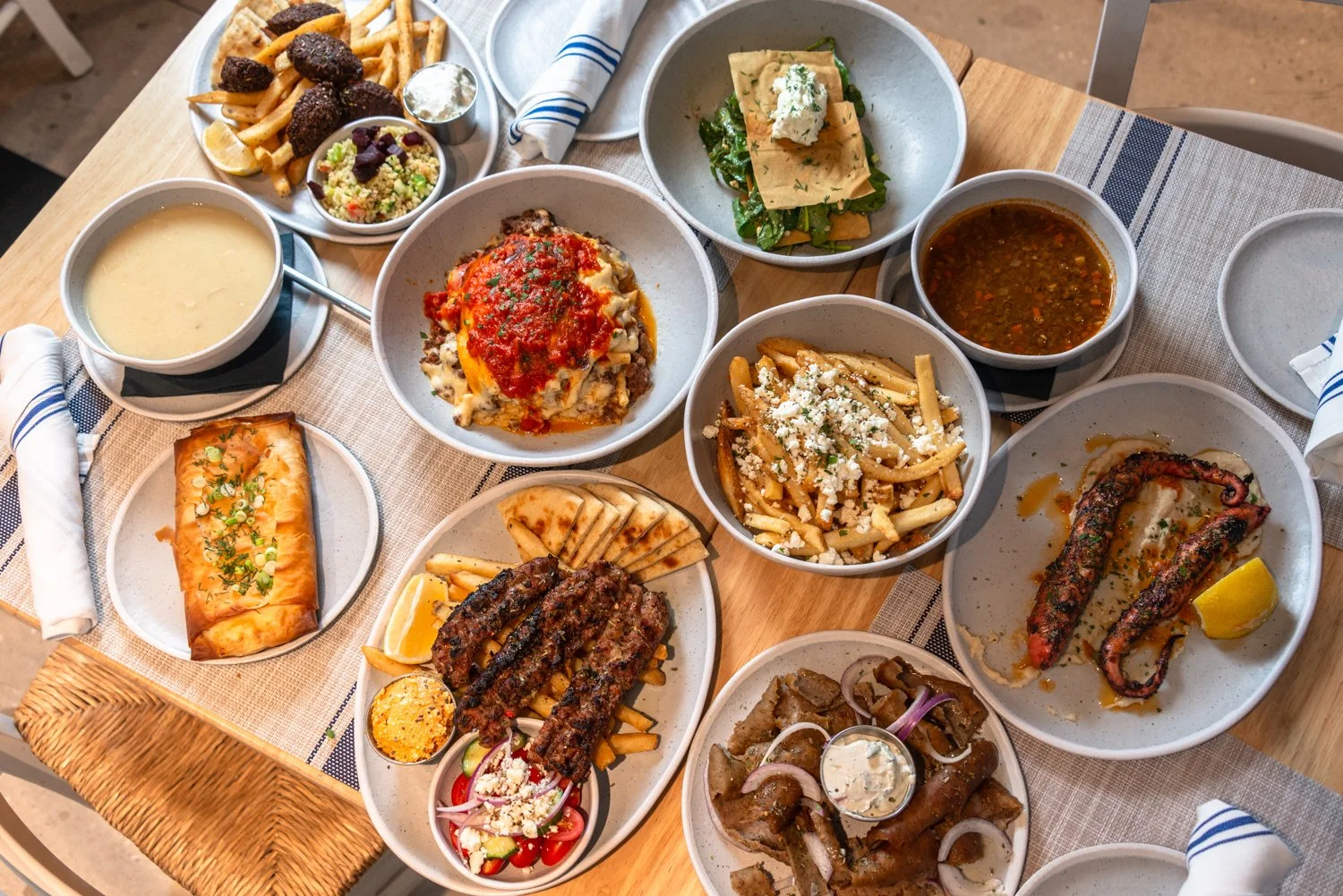 A variety of Greek dishes on a wooden table, including pasta dishes, grilled meats, salads, bread, and soups.