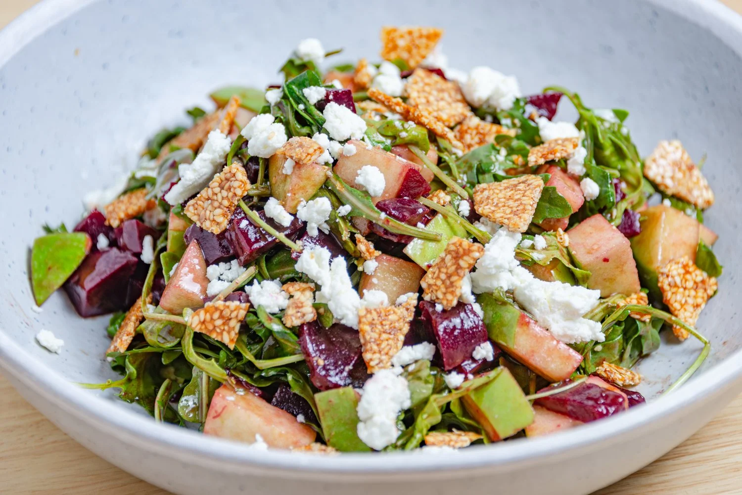A bowl of mixed greens salad with chopped apples, beets, crumbled cheese, and crispy rice crackers.