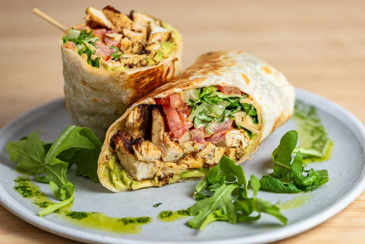 A plate with chicken wraps, one cut in half to show grilled chicken, lettuce, tomatoes, and other ingredients, garnished with fresh greens
