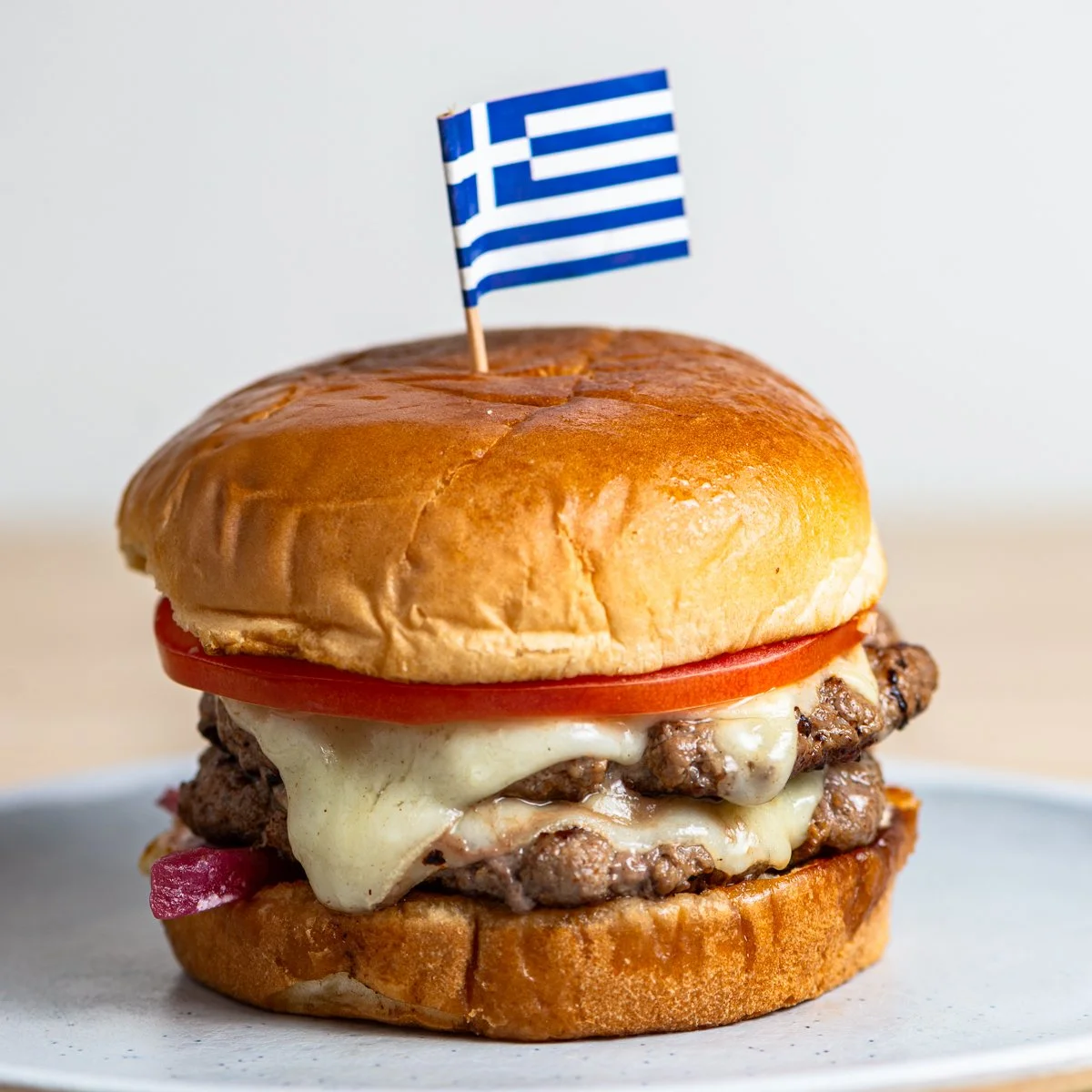 Close-up of a lamb burger with cheese, tomato, onions, and a Greek flag on top.