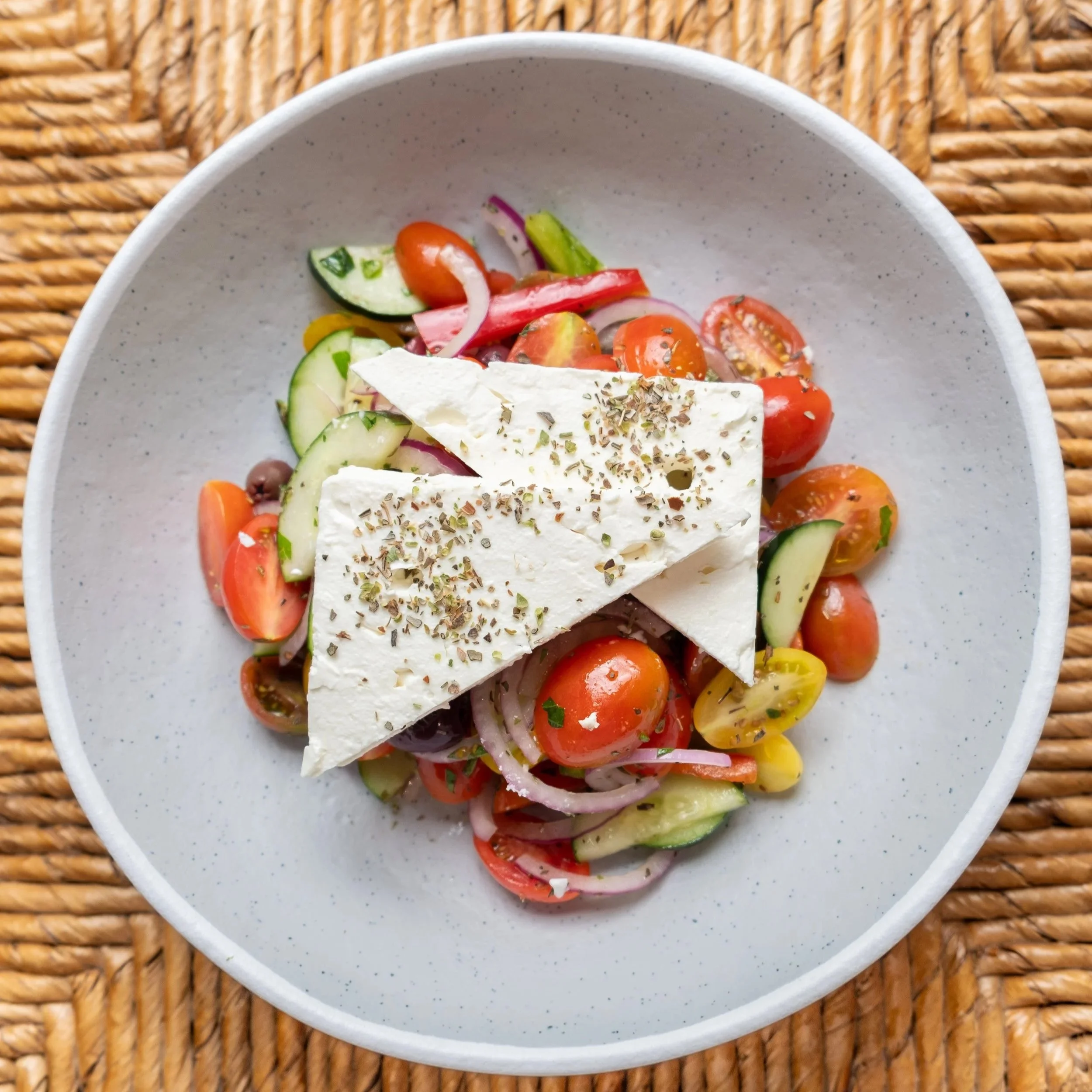 A bowl with a colorful Greek salad containing cherry tomatoes, cucumber, red onions, and basil, topped with slices of feta cheese and herbs, placed on a woven placemat.