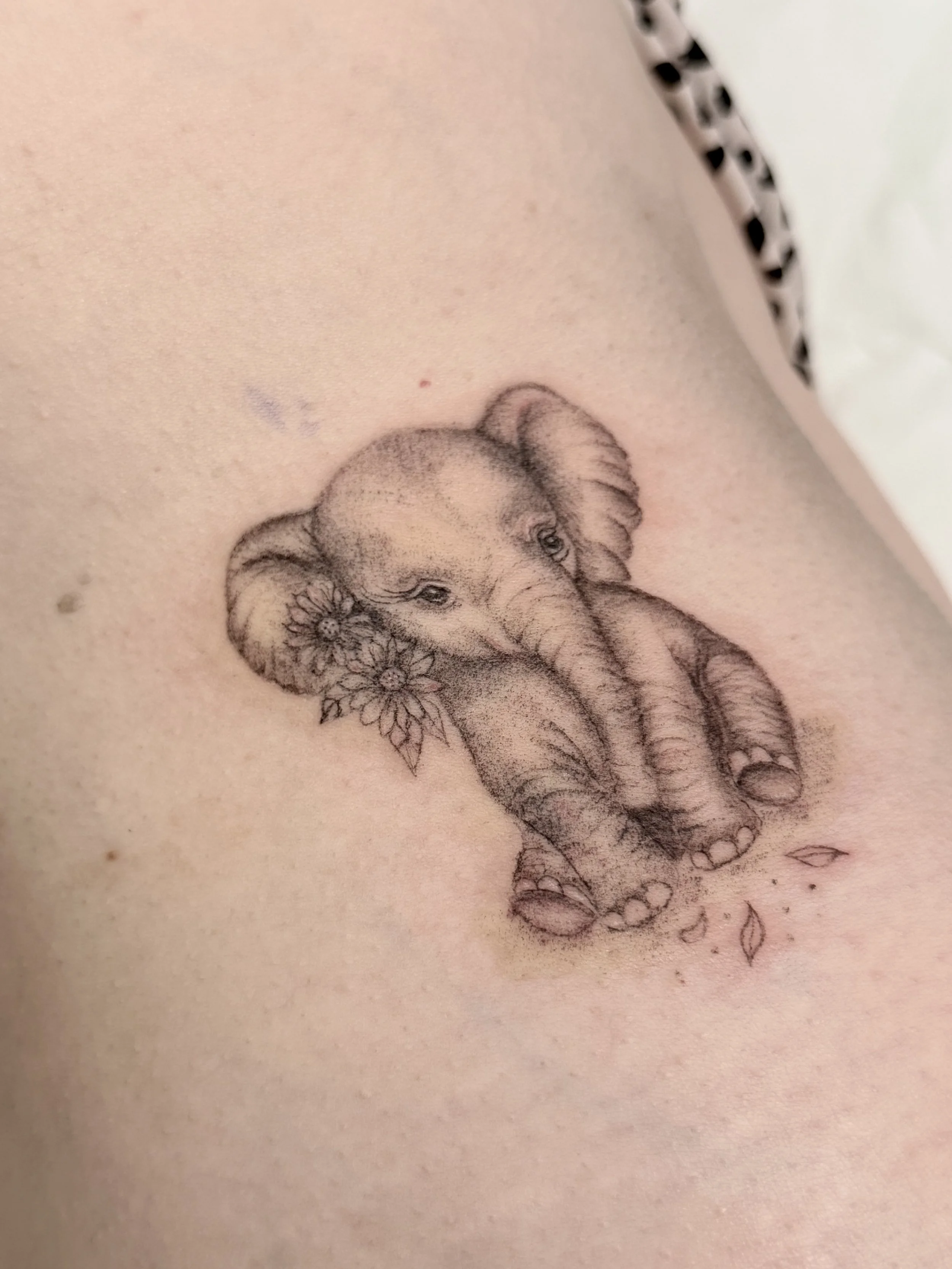 Tattoo of a baby elephant with flowers on its ear, sitting on the skin.