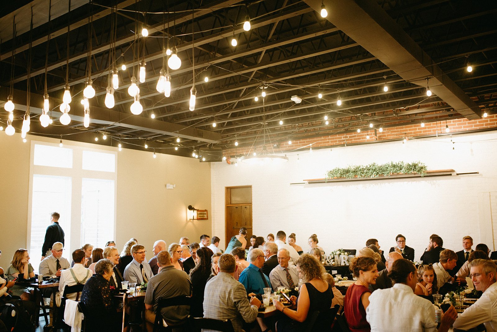 Edison bulb lights hang beneath exposed steel trusses and red brick at a wedding reception inside 8th & Main.
