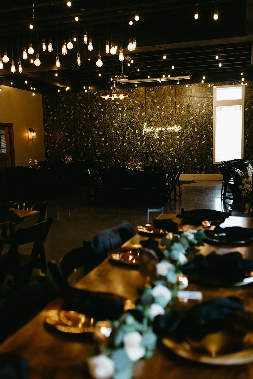 The reception hall’s deep-green botanical wall at 8th & Main, with a neon “love you more” sign and Edison bulbs overhead.