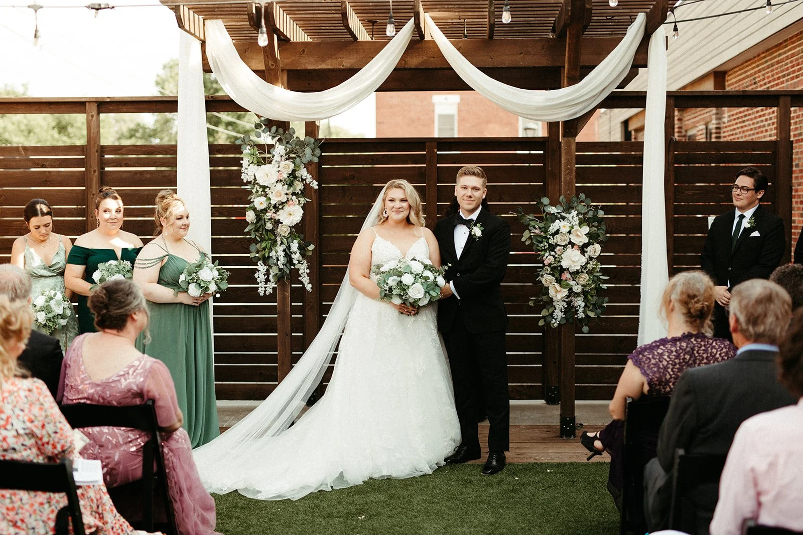 A bride and groom stand at the draped timber pergola in the 8th & Main courtyard, white florals climbing the posts.