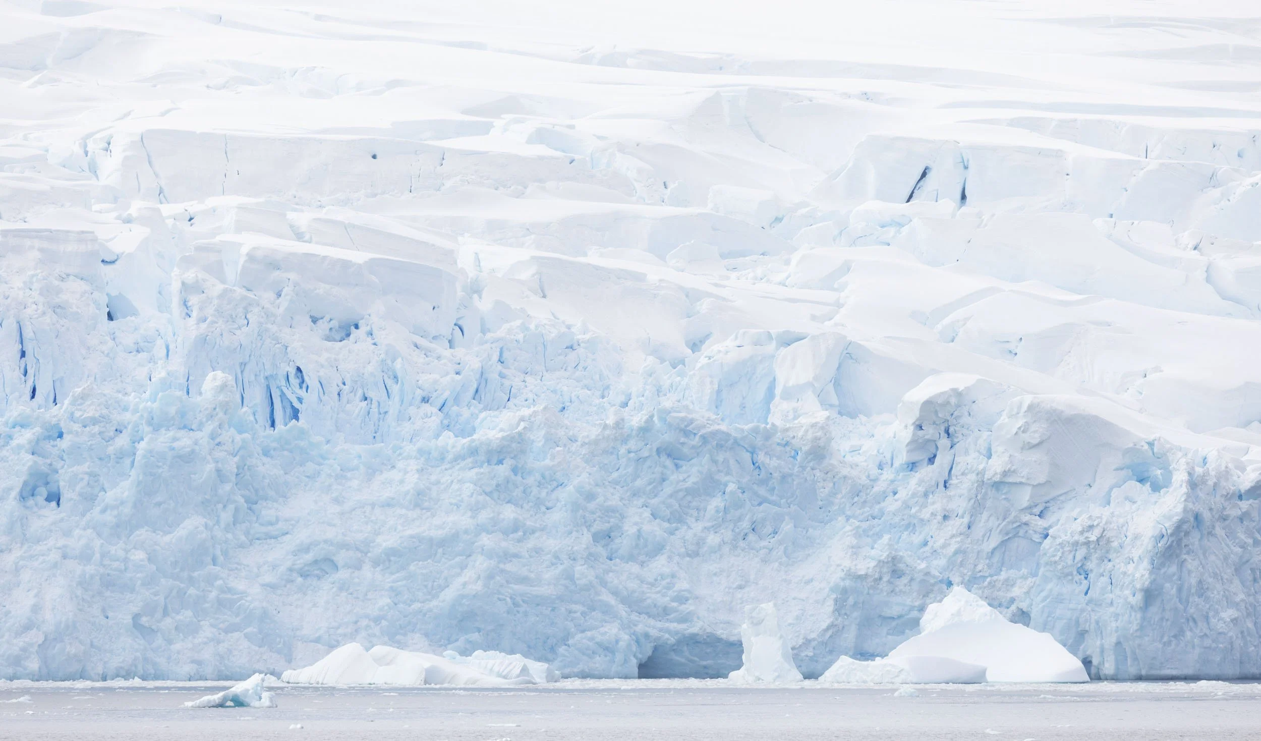 The blue and white hues of the face of a glacier fill the frame, creating interesting abstract textures and patterns throughout the image. A tiny strip of water is visible in the foreground with bits of calved ice floating by. Taken in Antarctica.