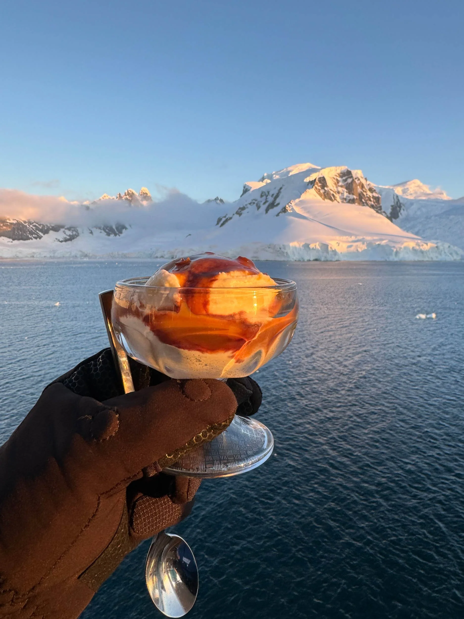 A gloved hand holds up a spoon and a glass bowl of vanilla ice cream drizzled with hot fudge in front of a soft sunset drenched mountain range and glacier across the sea in Antarctica.