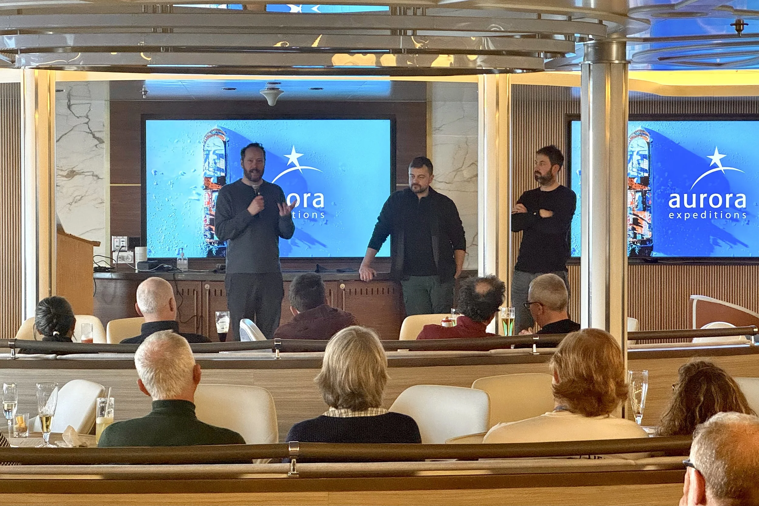 Nigel Danson, Mads Peter Iversen, and James Popsys stand and address a room full of people aboard the Aurora Expeditions ship the Sylvia Earle in Antarctica.
