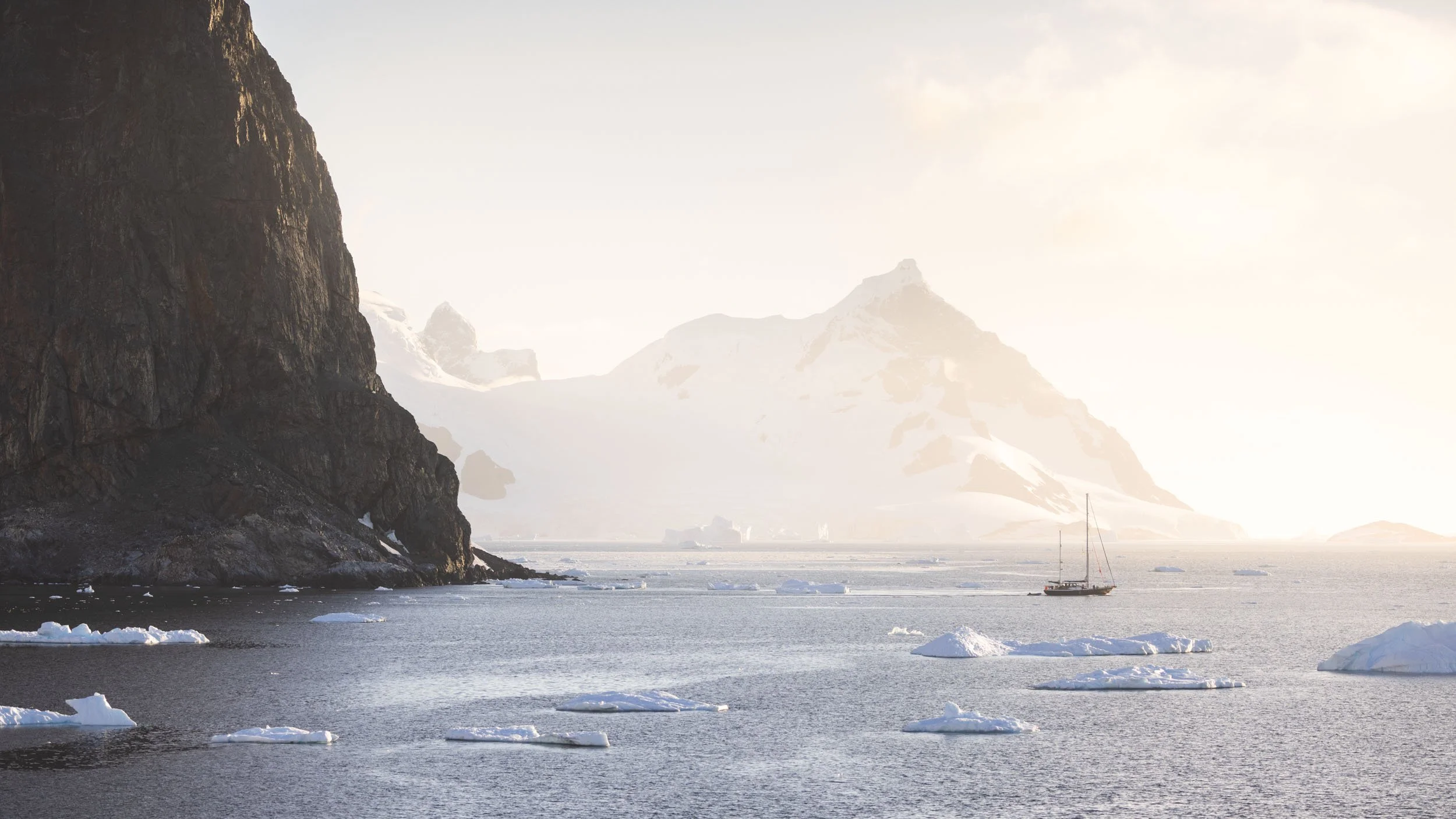 A lone sailboat passes through the expansive, iceberg-riddled Antarctic waters as soft, warm sunlight illuminates the gargantuan mountains of the Antarctic Peninsula. Captured in February 2024 from the decks of the Aurora Expeditions Sylvia Earle.