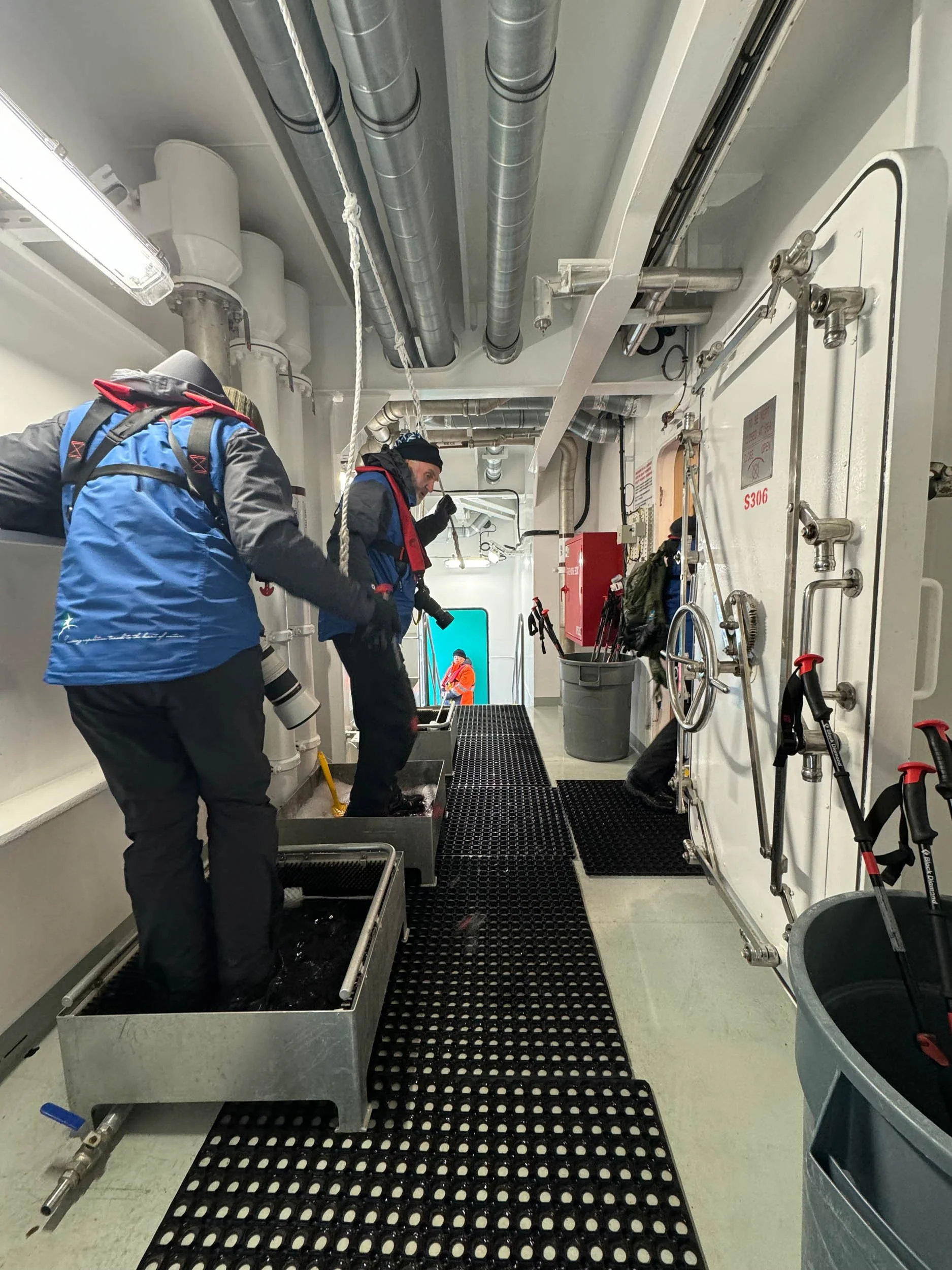 Two photographers dressed in full expedition gear, cameras dangling by their sides, step into metal basins to sanitize their  boots as they hold on to ropes for support, while aboard the inner decks of the Sylvia Earle expedition ship in Antarctica.