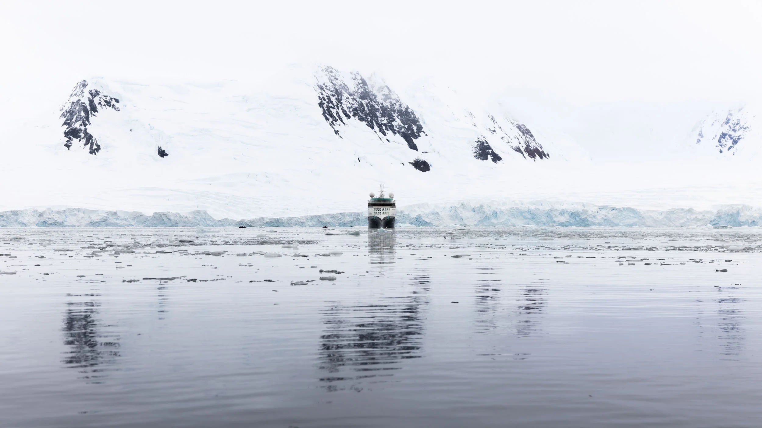 The Aurora Expeditions ship Sylvia Earle positioned in the distance facing the camera nose-on and exactly center in the frame, reflective water below, and endless glacier and mountain extend upwards and across. Taken in Borgen Bay, Antarctica.
