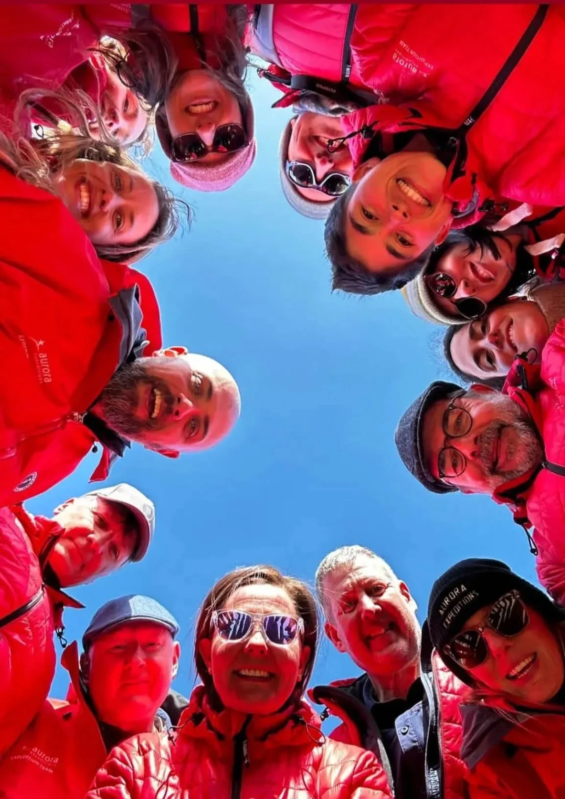 Fourteen smiling people dressed in red, Expedition Team members aboard Aurora Expeditions' Sylvia Earle ship, surround the frame from all sides looking in, as the camera points to the sky in Antarctica.