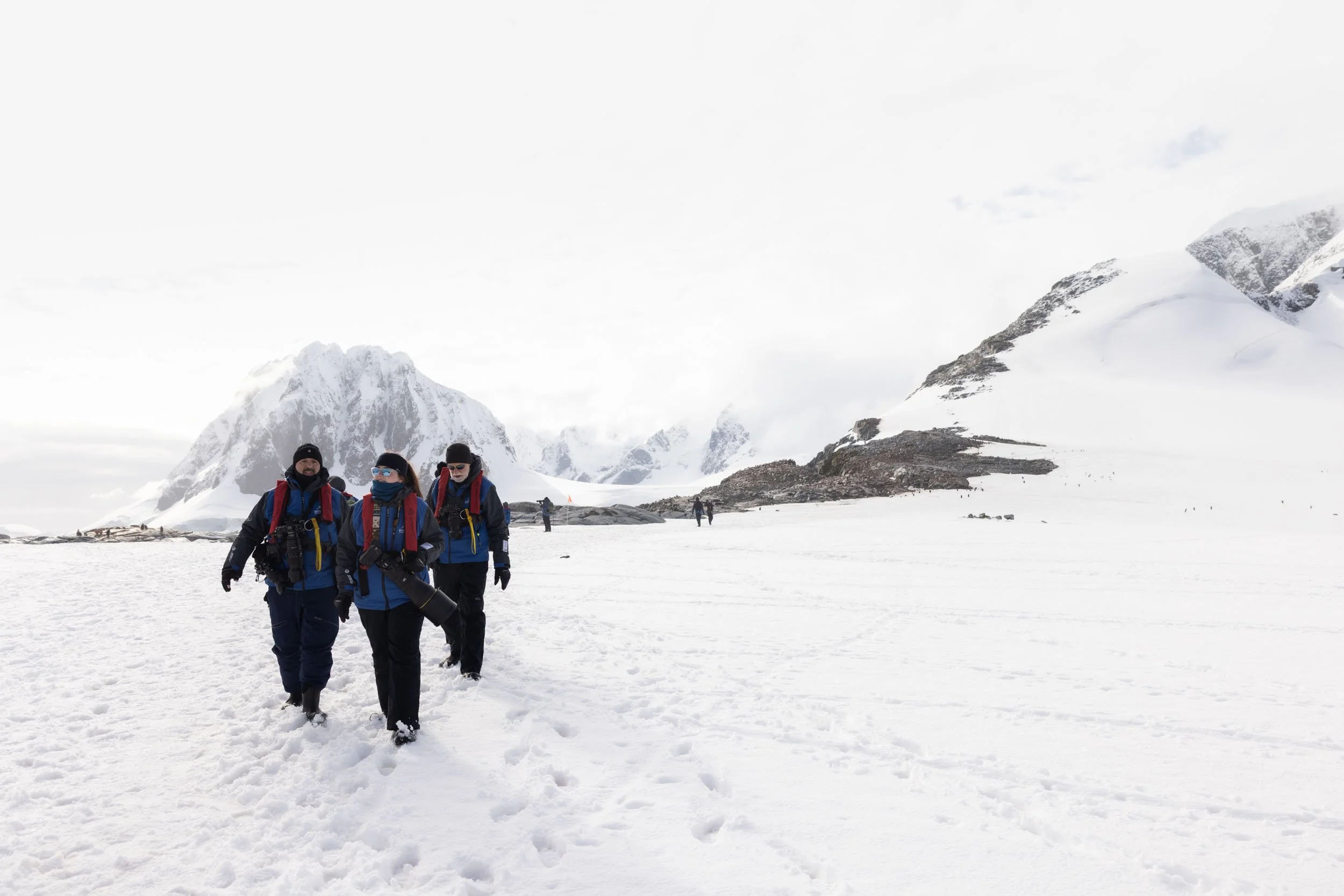 Rachel Bigsby, outfitted in full expedition gear and holing a large camera, leads two other photographers as they walk through a vast dramatic snow covered landscape near Port Charcot, Antarctica.