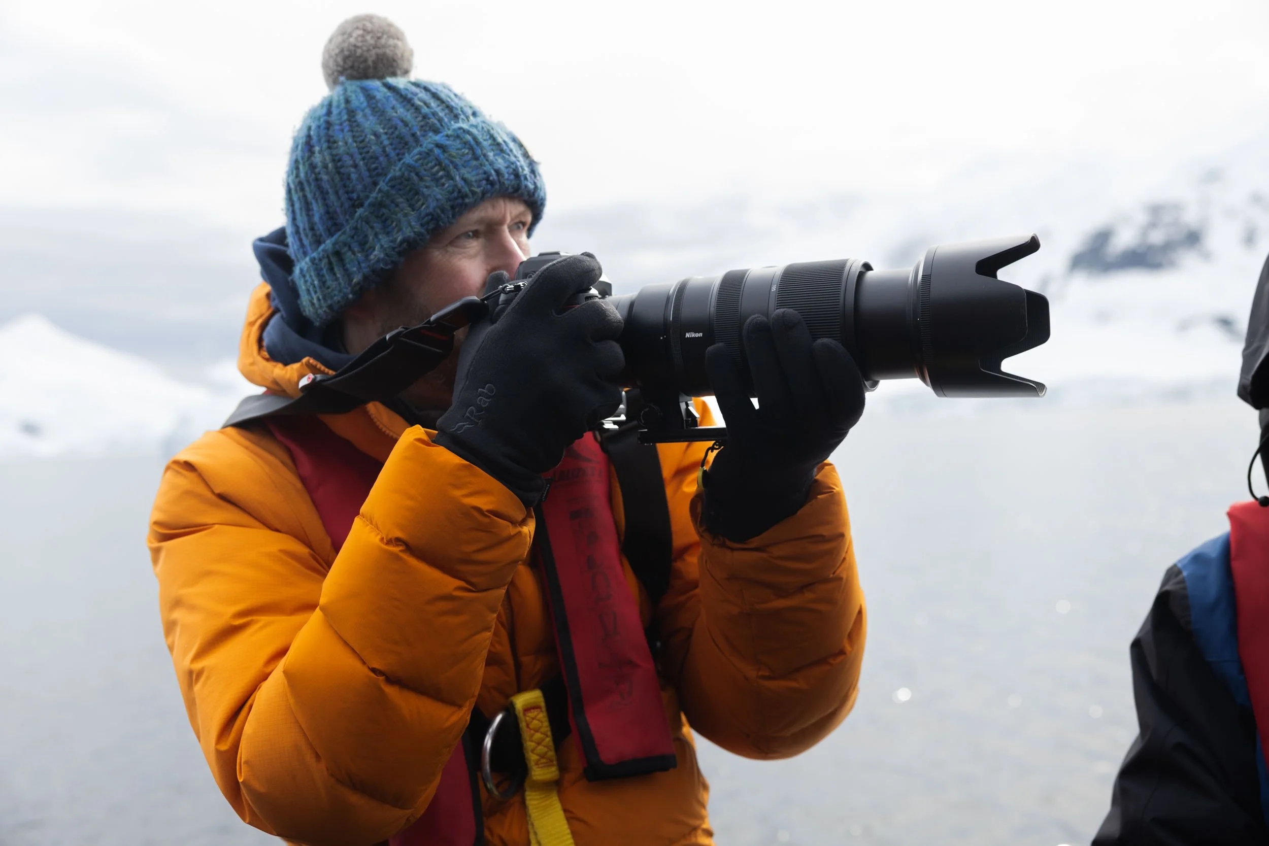 A profile portrait of Nigel Danson in a zodiac looking past his long lens camera to spot his subject and line up the shot. Taken in Skontorp Cove, Antarctica.