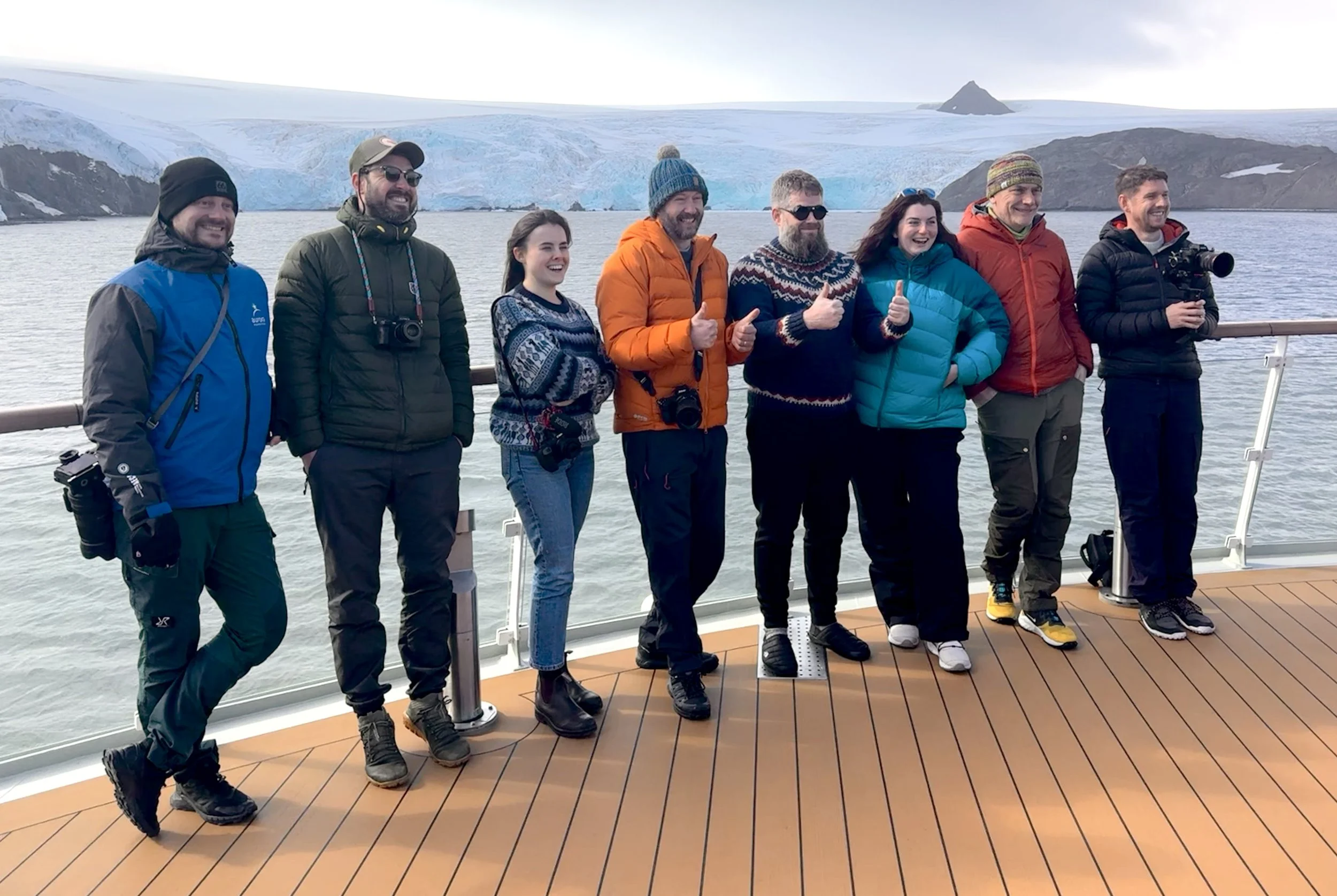 Mads Peter Iversen, James Popsys, Dani Connor, Nigel Danson, Jeroen Van Nieuwenhove, Rachel Bigsby, Adam Gibbs, and Rick Bebbington stand on the deck of the Sylvia Earle in Antarctica and smile.