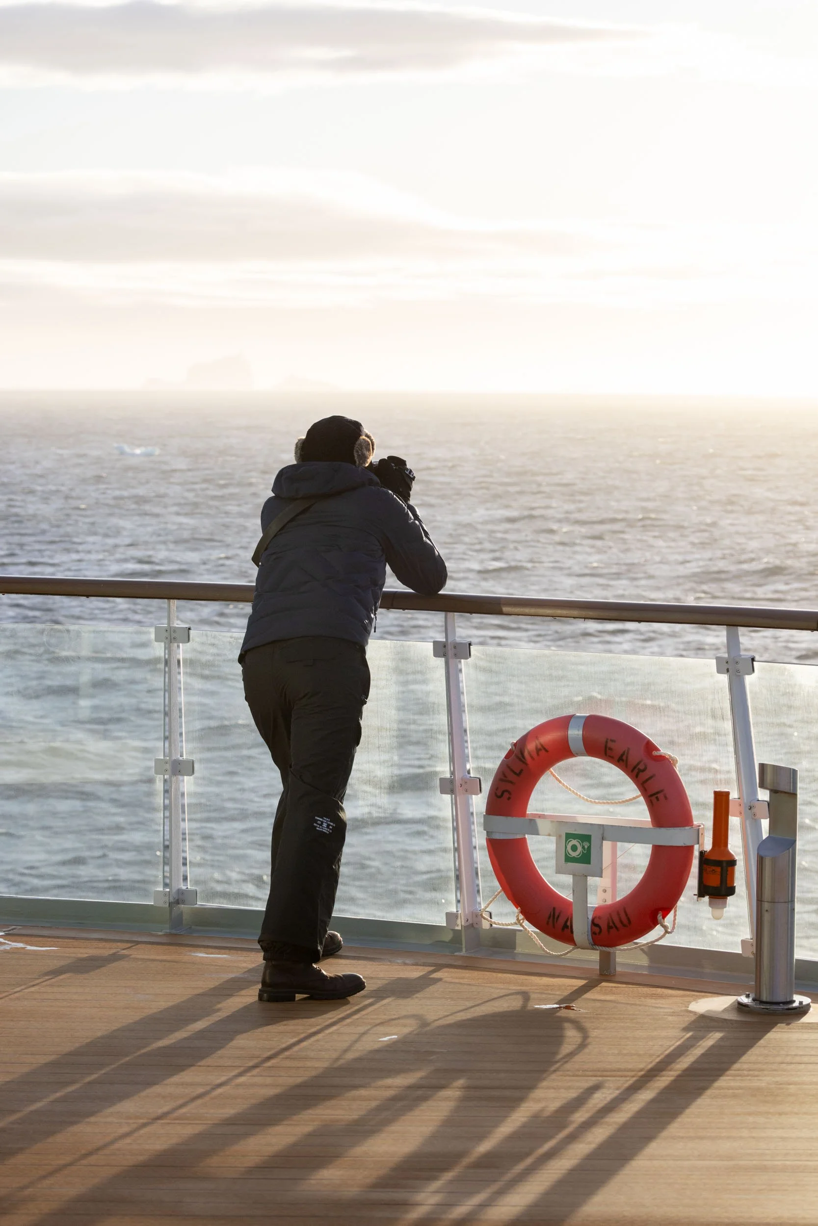 A photographer braces himself against the outer railing on the upper deck of the Sylvia Earle ship, camera in hand, carefully taking a shot looking out to sea, icebergs in the distance, as soft low-angle sunlight casts long shadows across the deck.