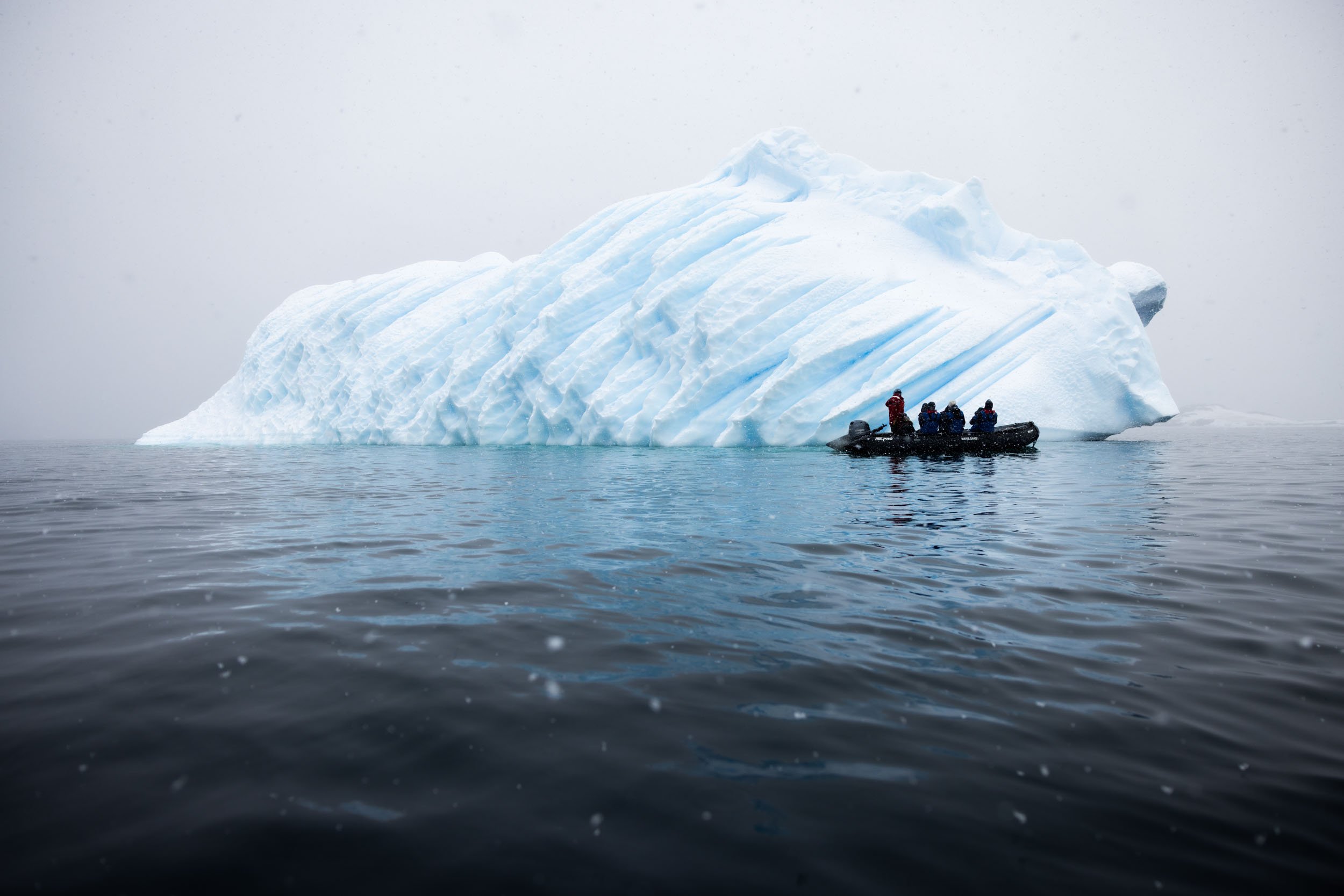 A zodiac in silhouette passes by a recently overturned iceberg on a snowy day in Recess Cove, Antarctica.