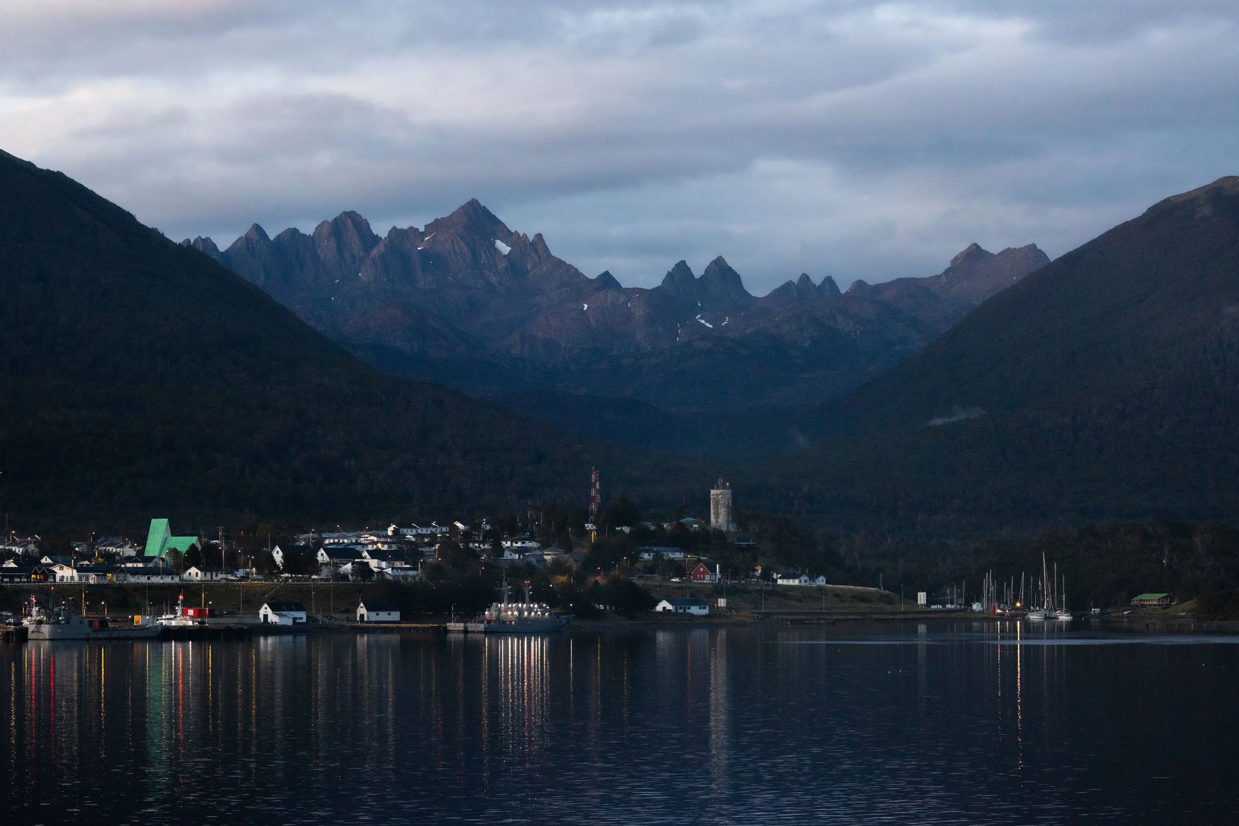Towering mountains of Patagonia frame the small port town of Puerto Williams, Chile, as soft blue-hour sunlight barely begins to illuminate the Beagle Channel while buildings and sailboats reflect in the calm water.