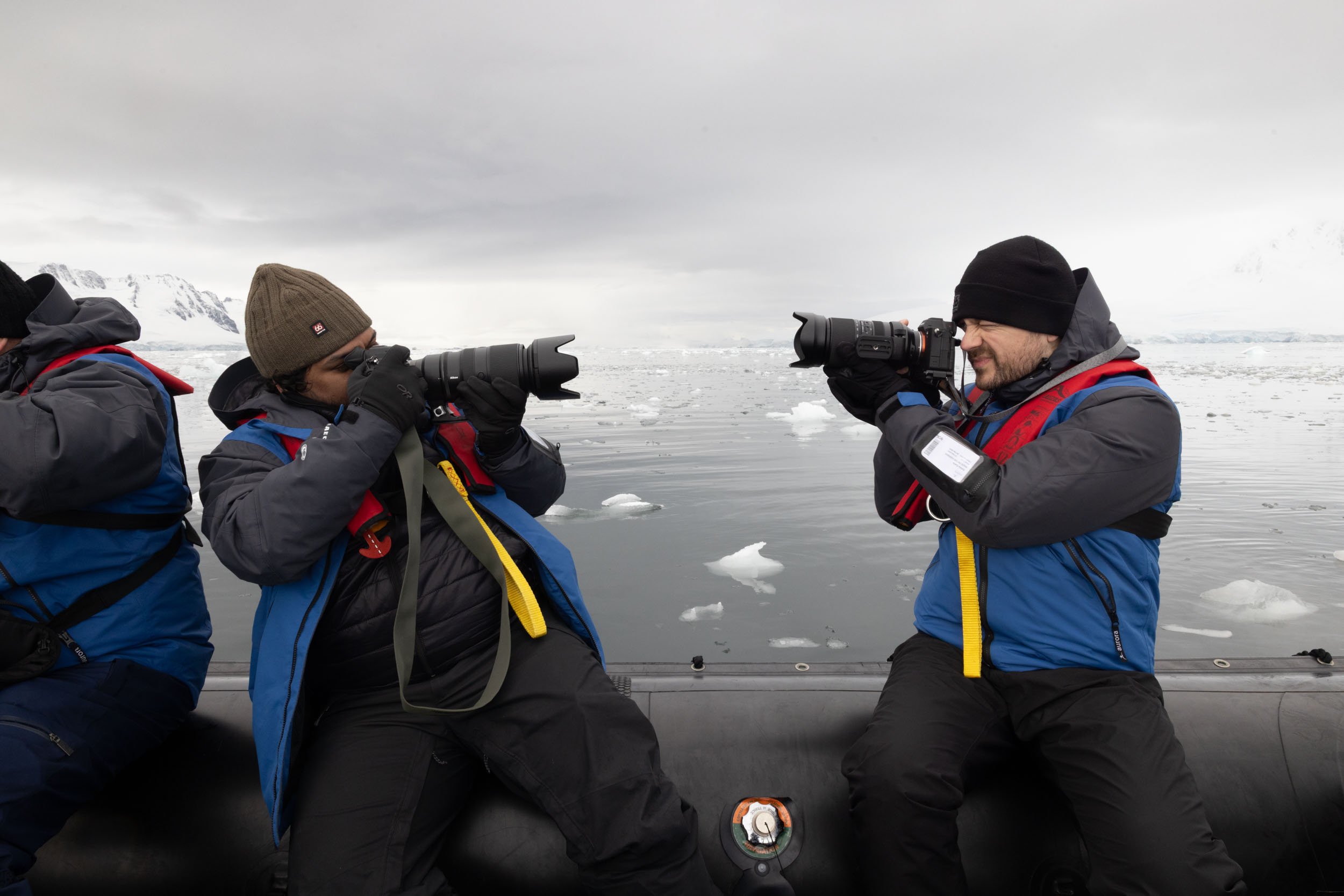 Mads Peter Iversen sitting in a zodiac, camera to his face, taking a picture of the person sitting directly next to him, while that person simultaneously takes a picture of Mads in a comical "camera battle" moment in Antarctica.