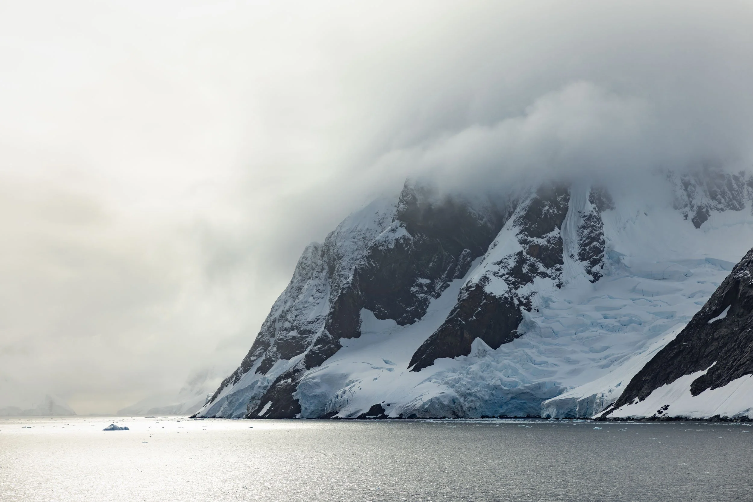 A massive mountain with a cascading glacier meeting the sea, overtaken by sunlight-drenched thick descending clouds, dominates the frame as a relatively tiny iceberg is juxtaposed in the water nearby in Antarctica.