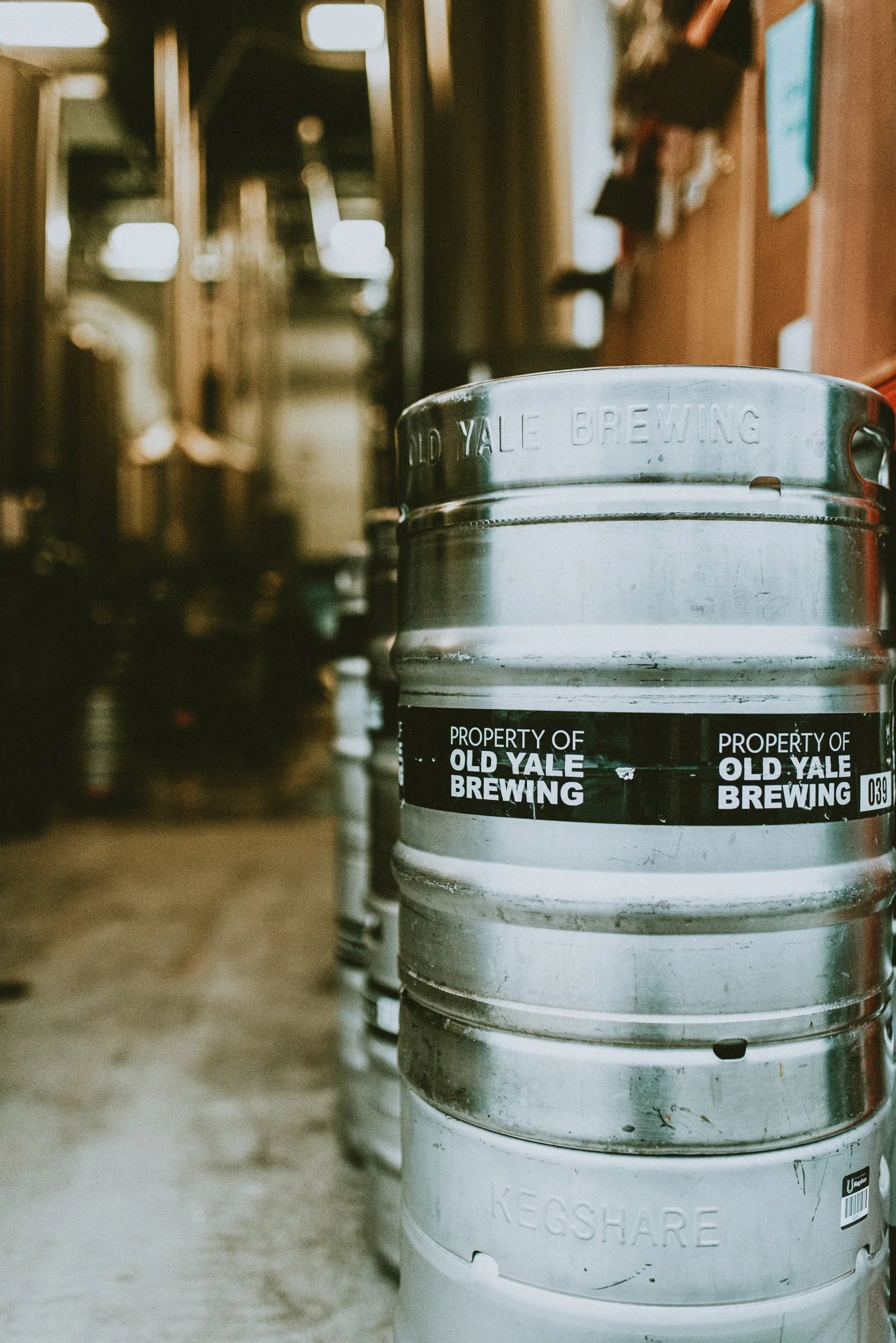 A row of stainless steel kegs in a brewery or taproom, with the front keg labeled 'KEGS HARE' and the front of the second keg stamped with 'OLD YALE BREWING'.