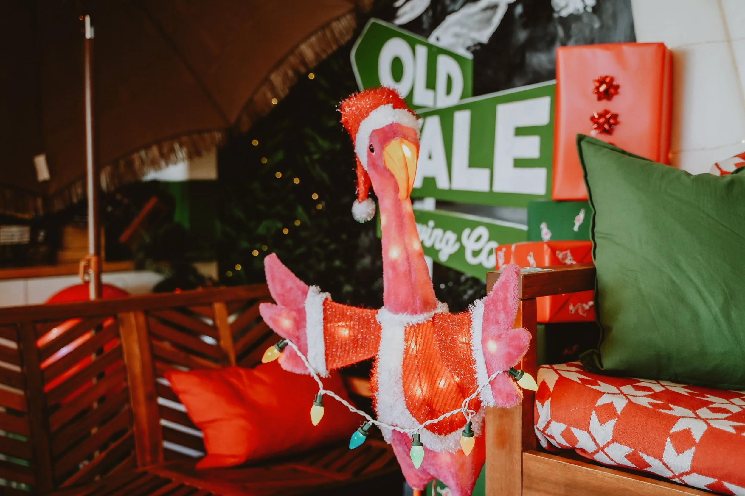 A festive display featuring a plush pink flamingo wearing a Santa hat, string lights, a green sign with white letters reading 'OLD SALE,' and wrapped Christmas presents including a red box with bows, all set on a wooden table with a green cushion and