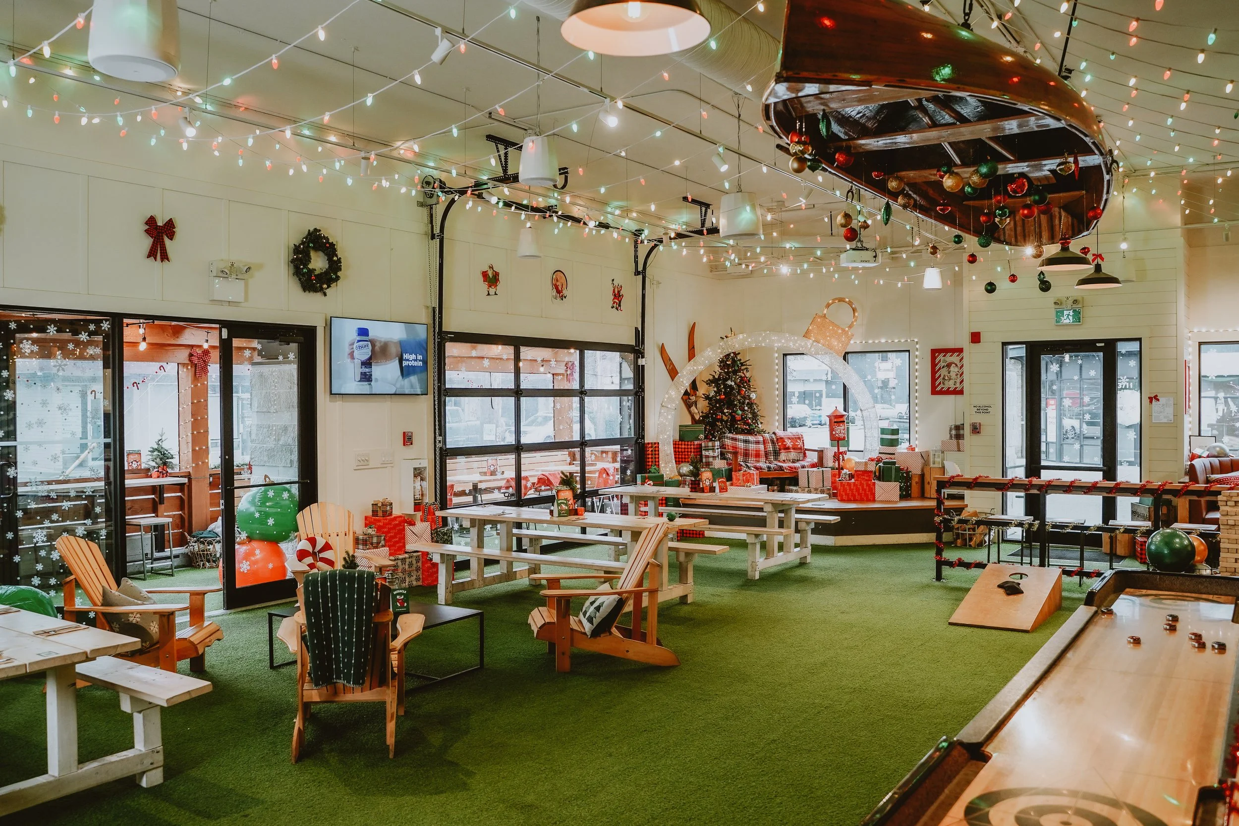 An indoor space decorated for Christmas with string lights, holiday wreaths, ornaments, a Christmas tree, and gift boxes. There are wooden chairs and tables, and a shuffleboard game on the floor. Windows have snowflake decals and outside snow can be 
