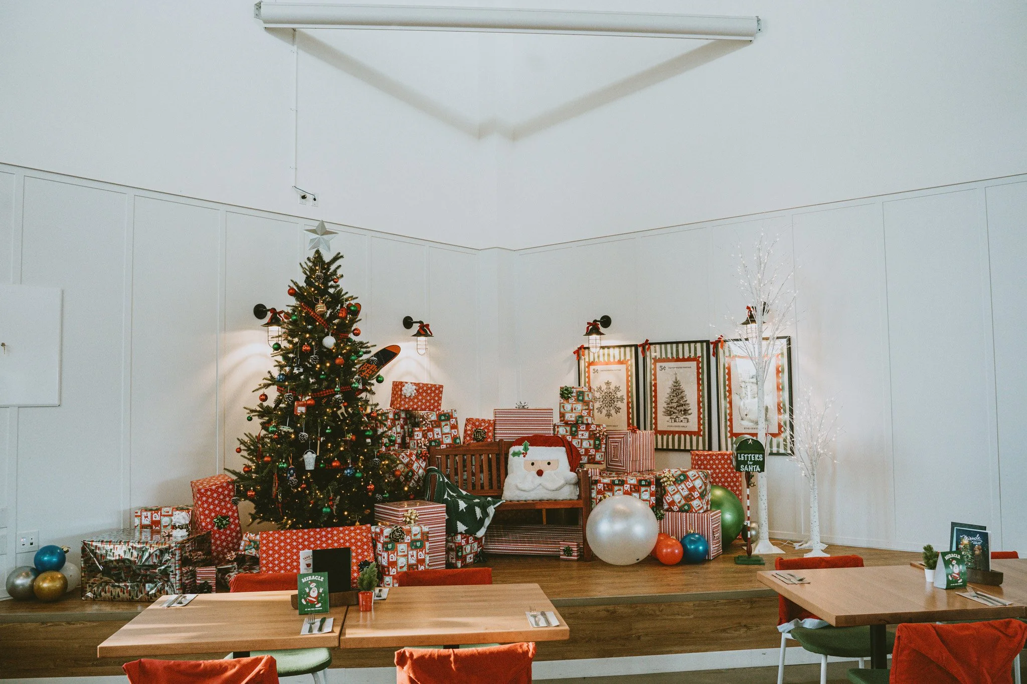 A decorated Christmas tree with presents underneath and festive decorations in a room with white walls, a string of lights, and holiday signs.