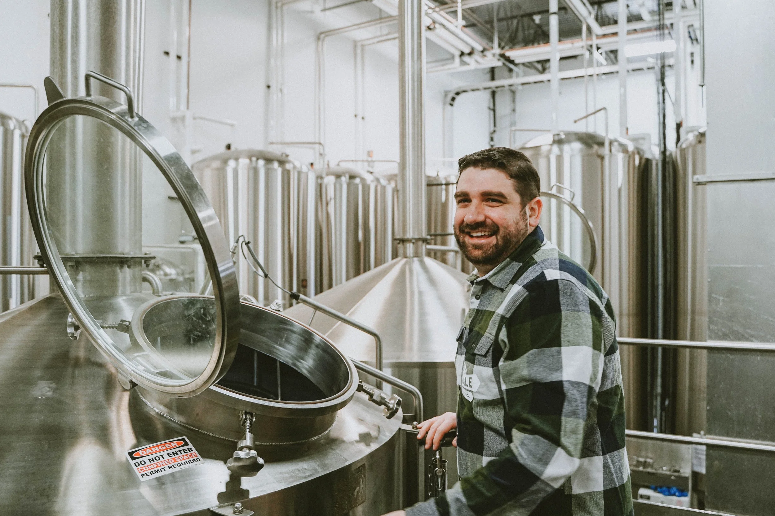 A man with a beard smiling while standing in a brewery, surrounded by large stainless steel brewing tanks.