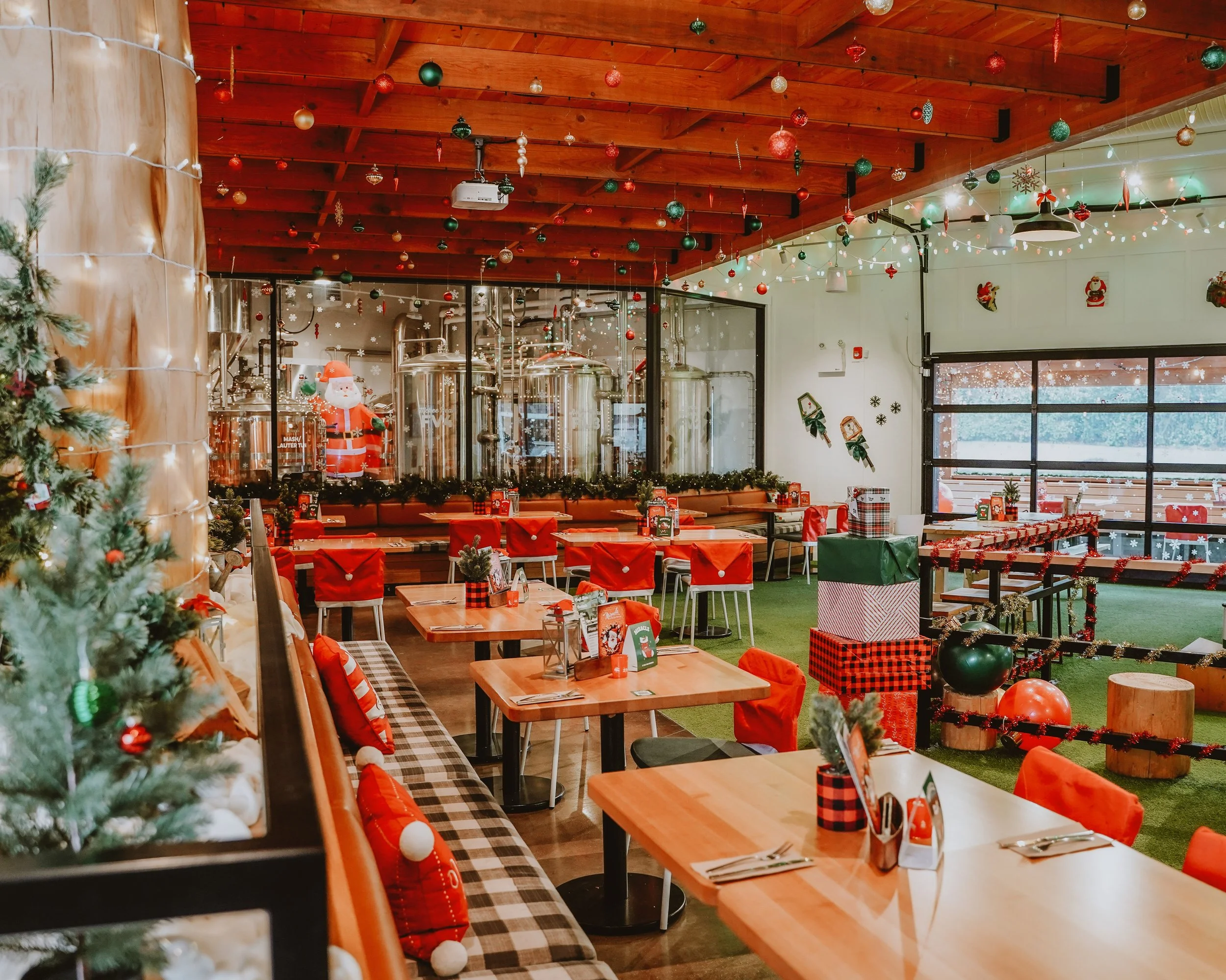 A festive indoor restaurant decorated for Christmas. The ceiling is wooden with ornaments hanging, and there are Christmas trees, wrapped presents, and holiday decorations throughout the space.